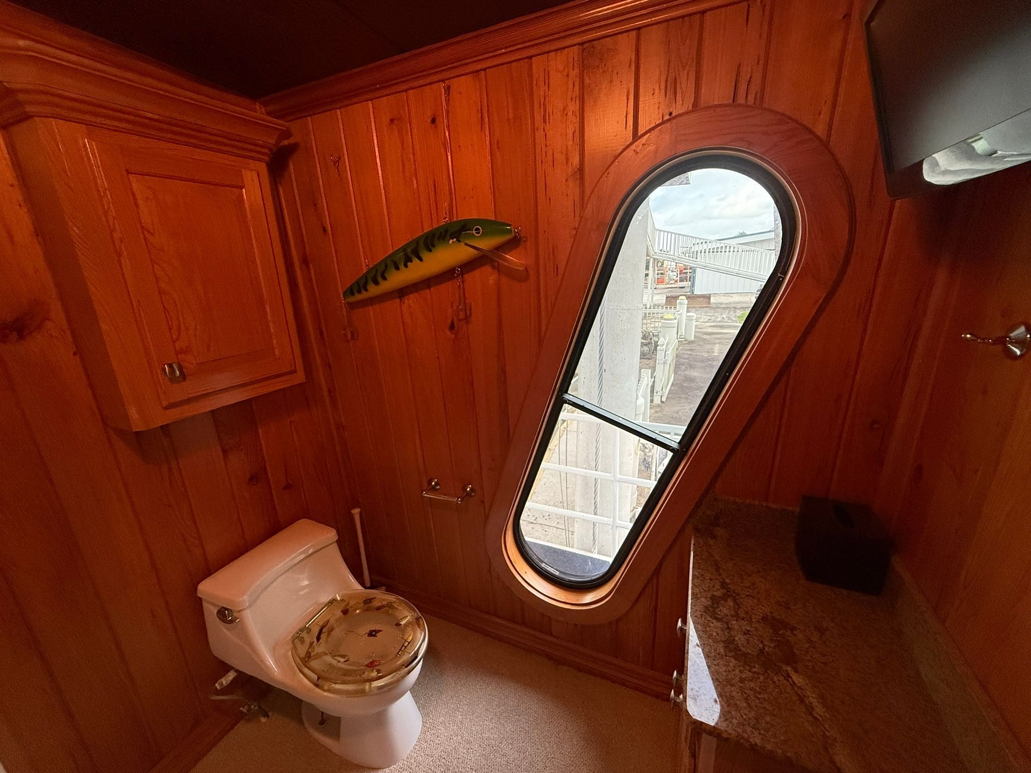 A bathroom with wood paneling, an oval window, and a toilet with fish decor.