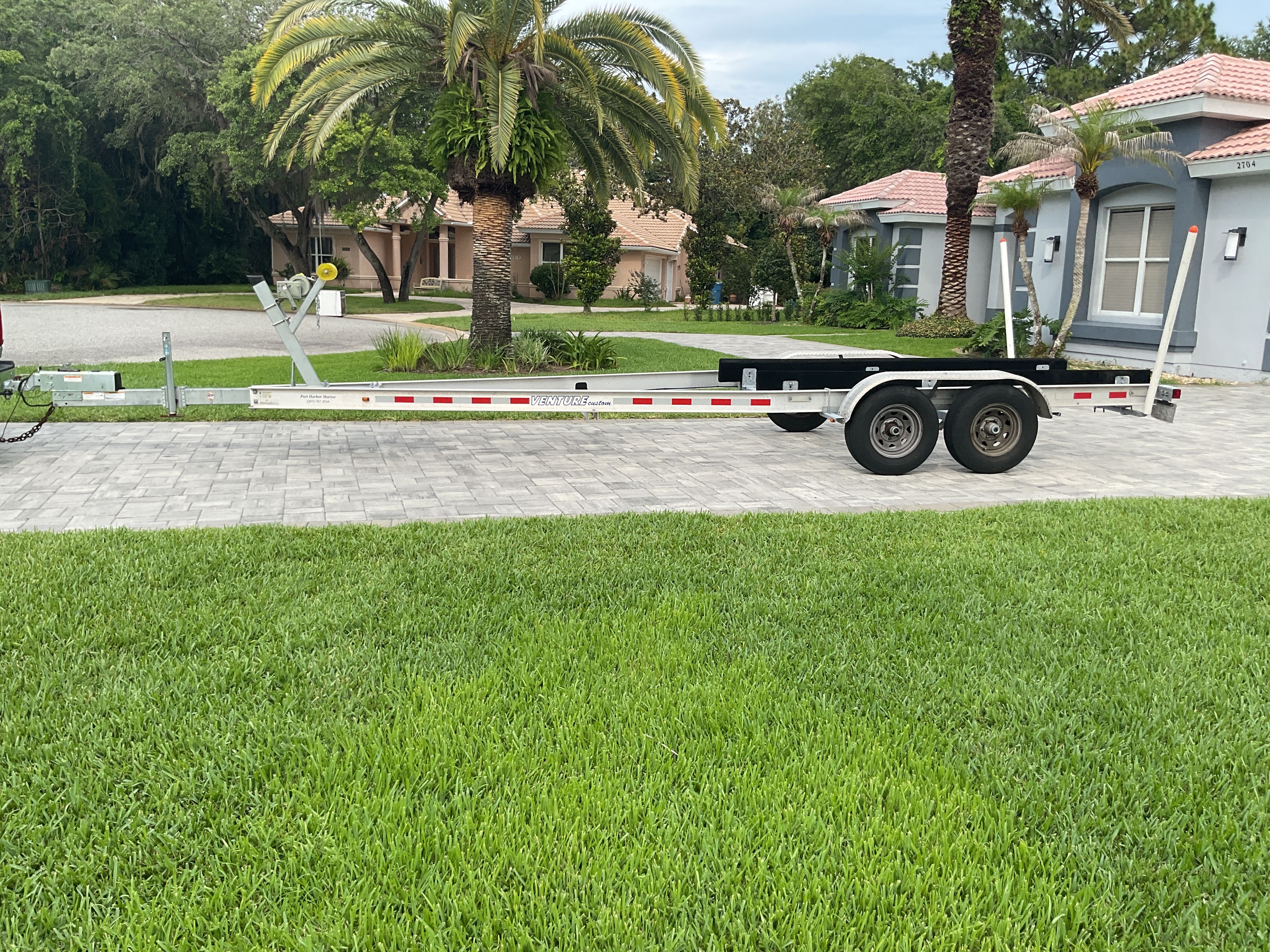 Boat trailer on paved driveway in front of houses and palm trees.