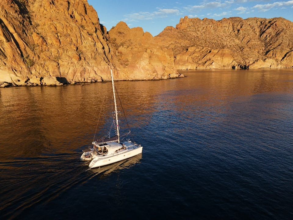 Catamaran sailing in a calm bay near rocky coastline at sunset.