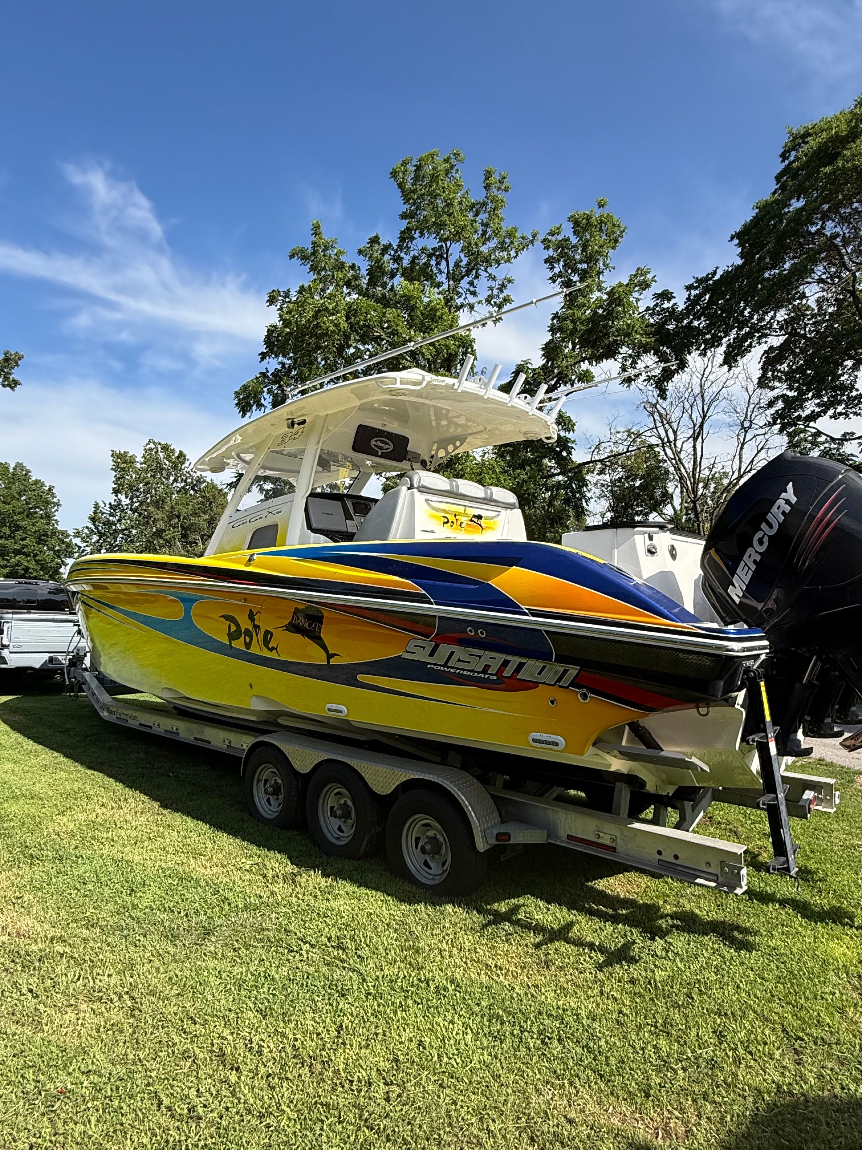 A colorful boat on a trailer on grass, with trees in the background.