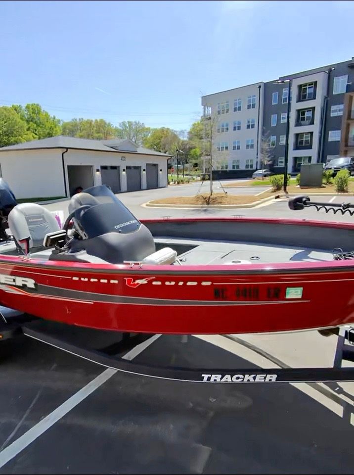 A red Tracker boat on a trailer in a parking lot, with modern buildings in the background.
