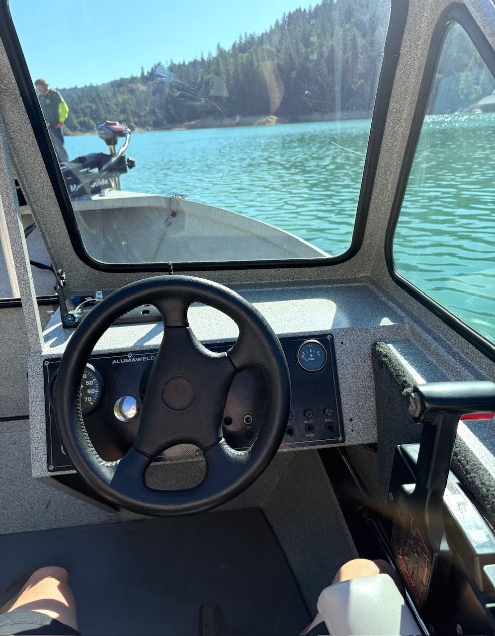 The image shows a boat's helm with a steering wheel and instruments. Water and a wooded shoreline are visible in the background.