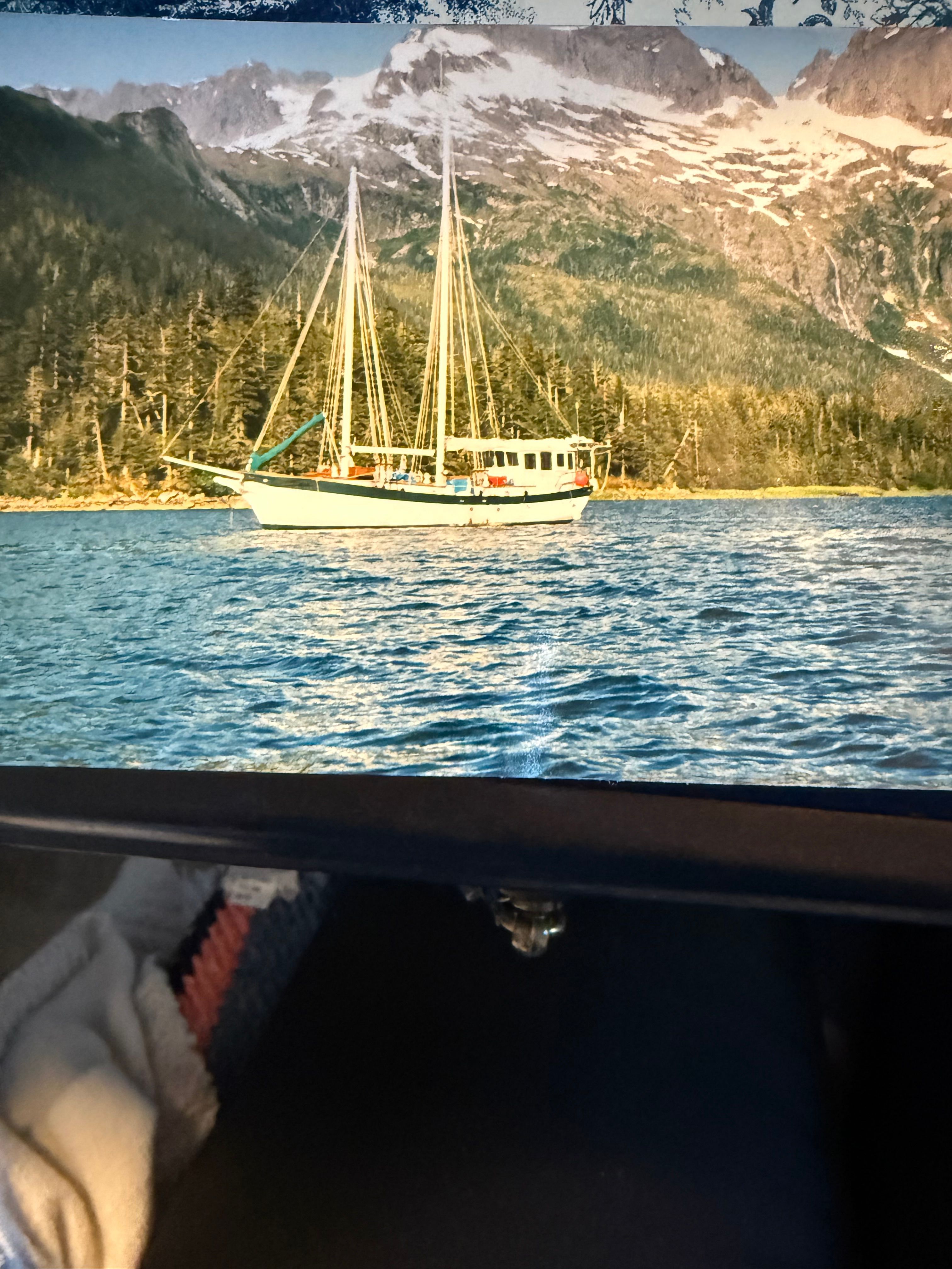 A 1981 schooner sails on calm water with a forested mountain backdrop.