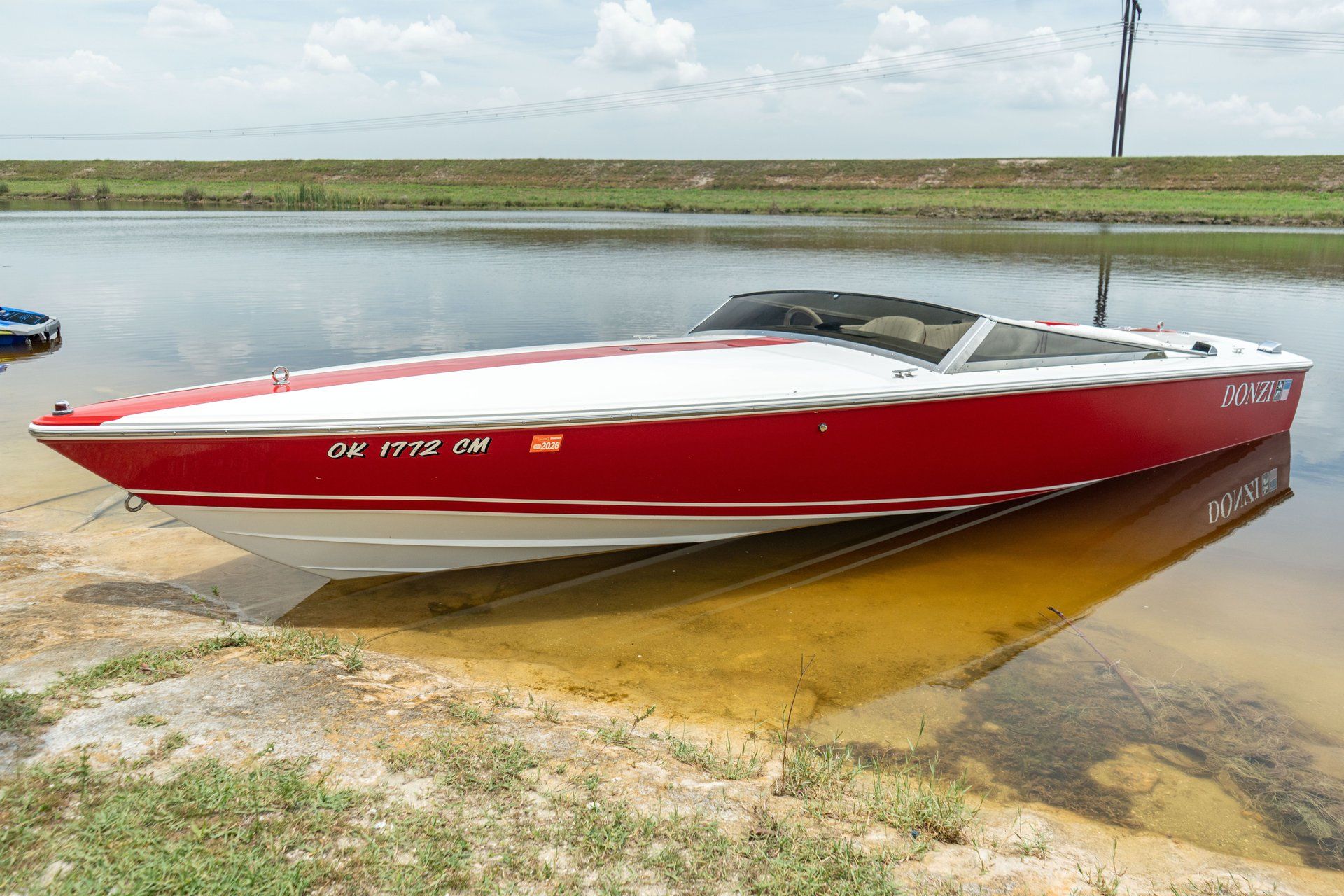 A red 1995 Donzi Classic boat is docked by the shore of a calm water body.