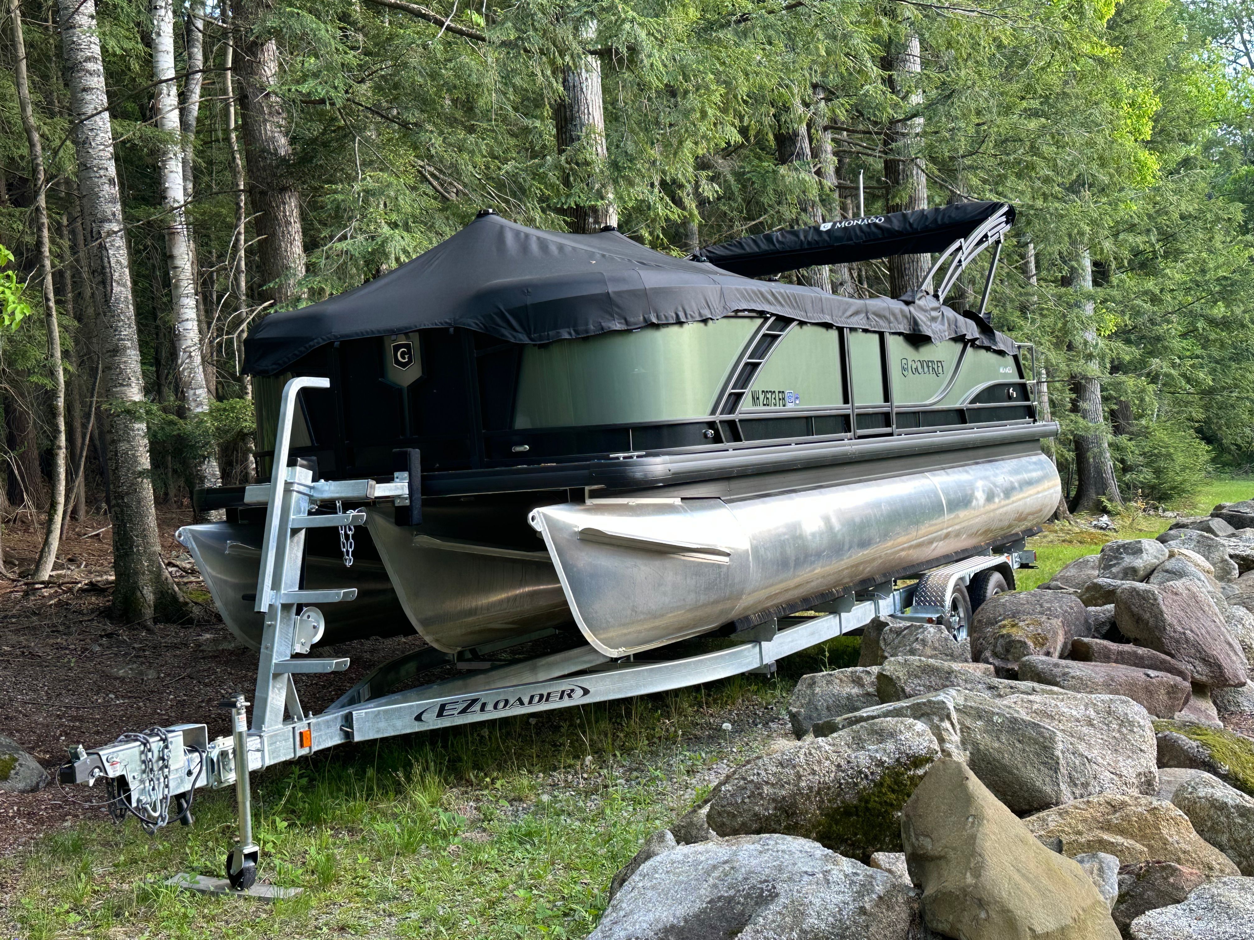 A 2025 Godfrey Monaco Cruise pontoon boat on a trailer in a wooded area.