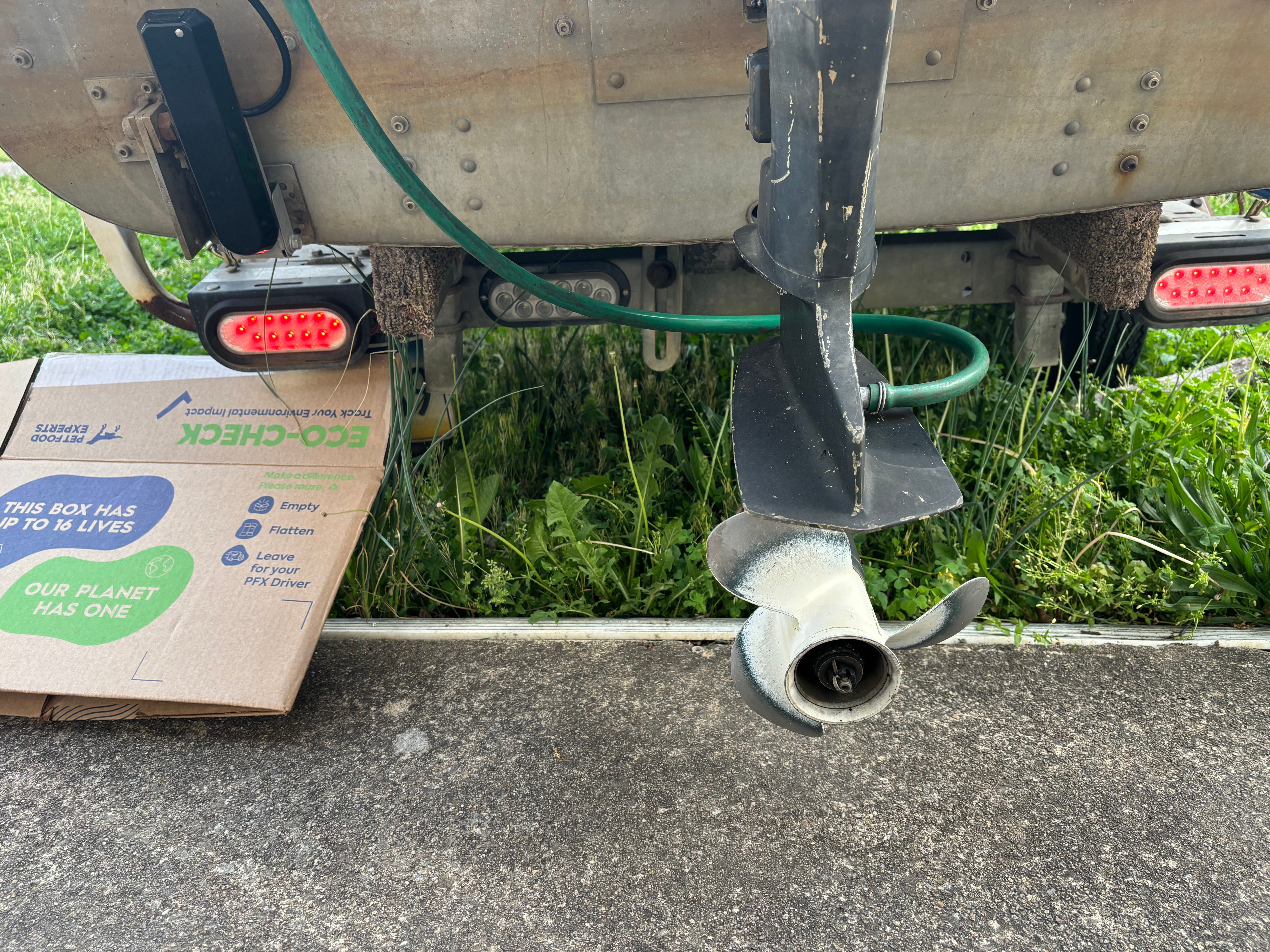 A boat motor above a concrete surface, surrounded by grass and a cardboard box.