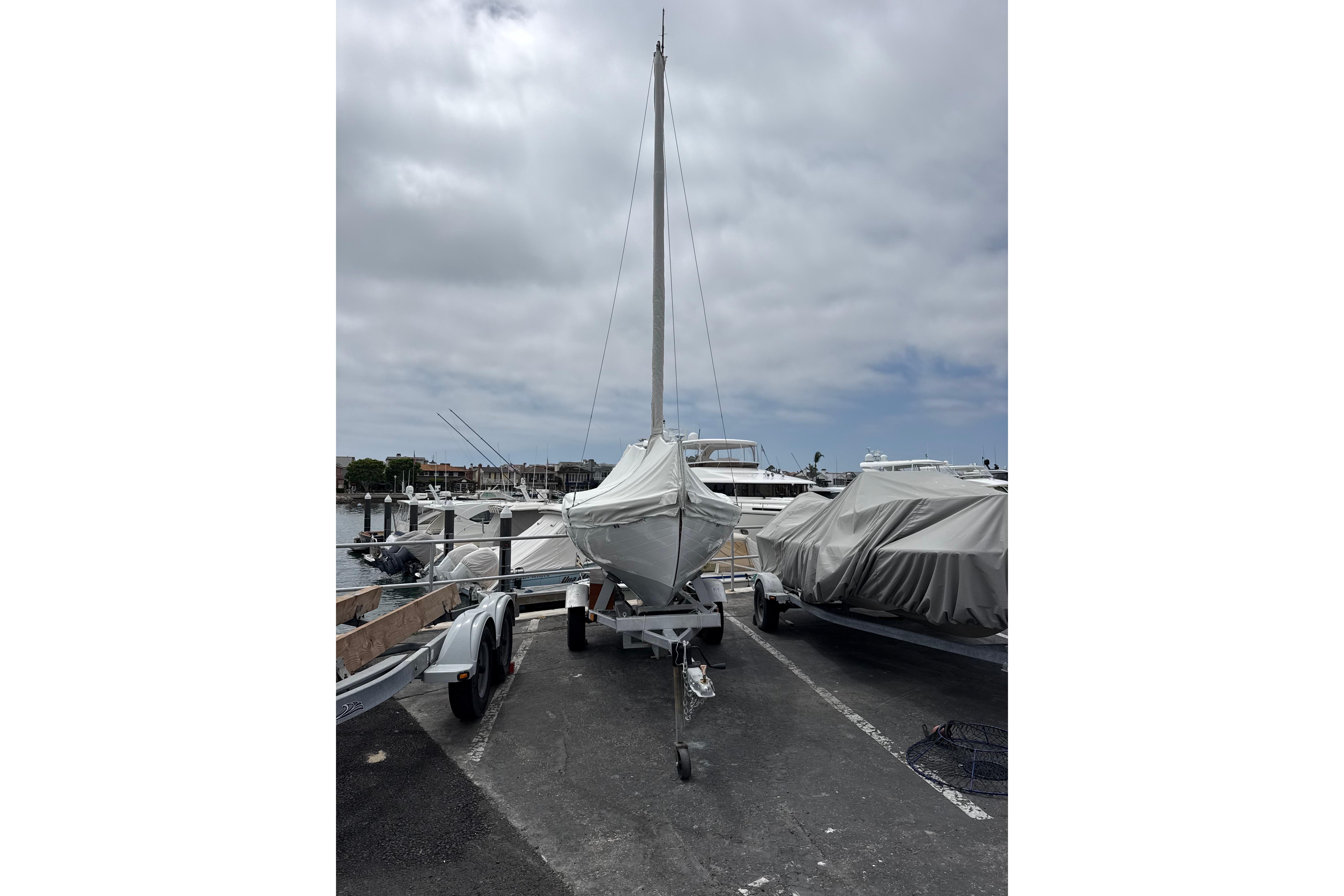 Covered 1920 Herreshoff 12.5 sailboat on trailer at marina under cloudy sky.