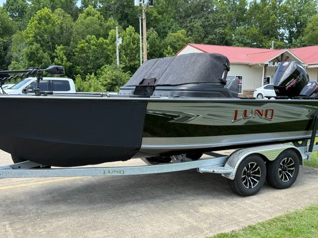 A 2025 Lund 2075 Impact XS boat on a trailer, surrounded by trees and buildings.