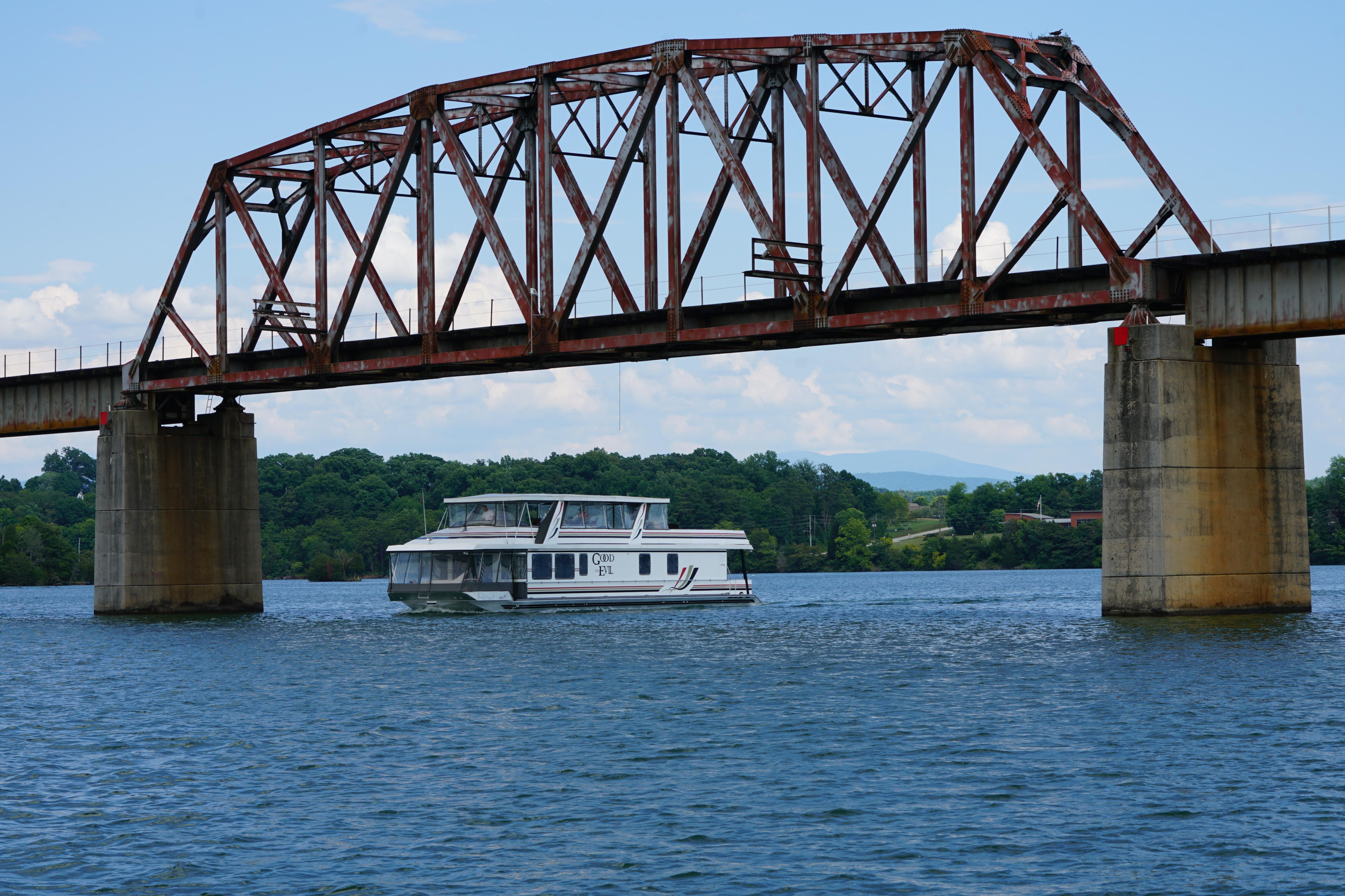 Houseboat cruising under a rusty bridge on a scenic lake.
