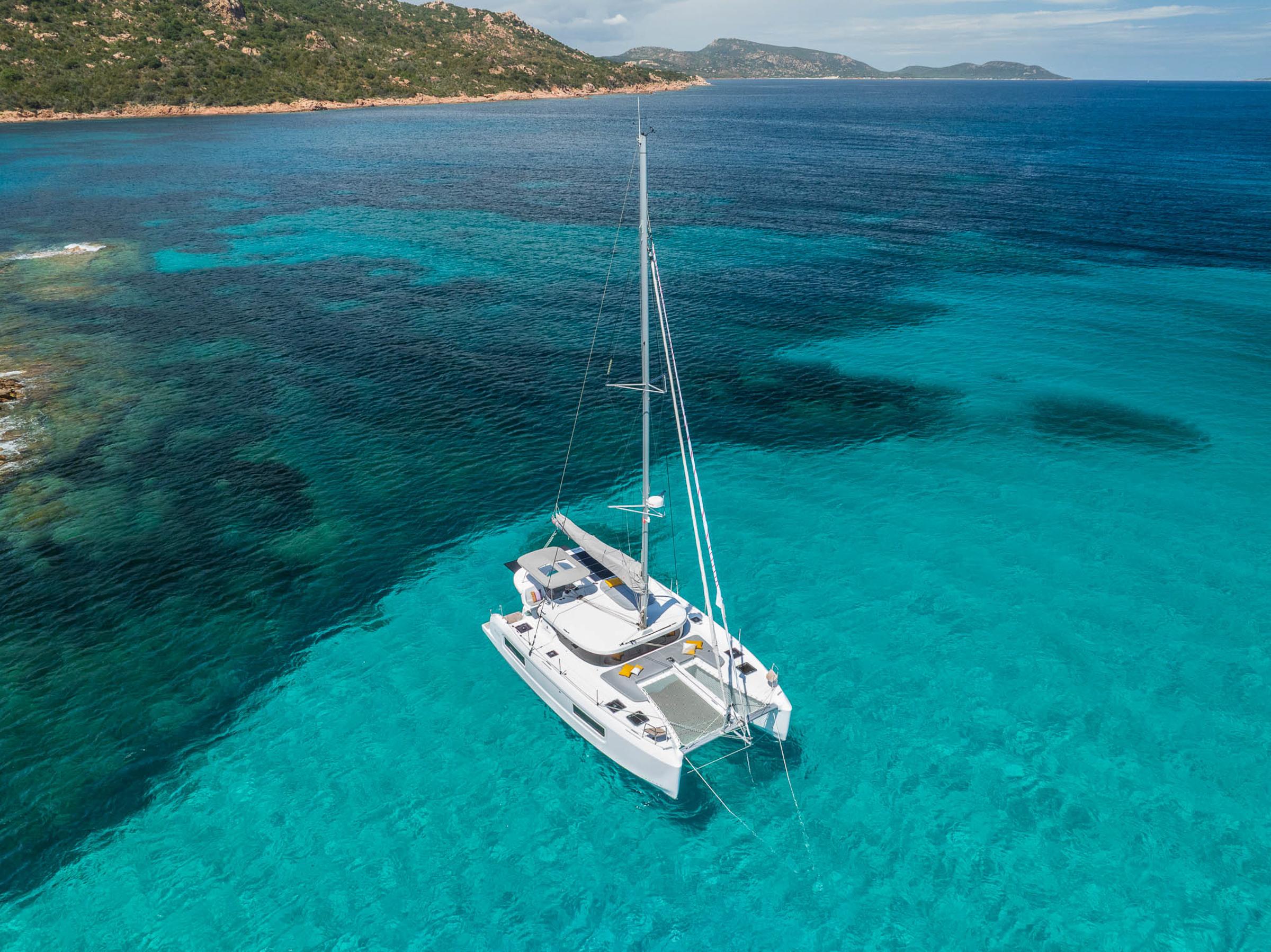 2025 Lagoon 38 catamaran sailing in clear turquoise waters near a rocky coastline.