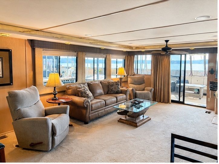 The interior of a houseboat with comfy armchairs, a sofa, and large windows overlooking the water.