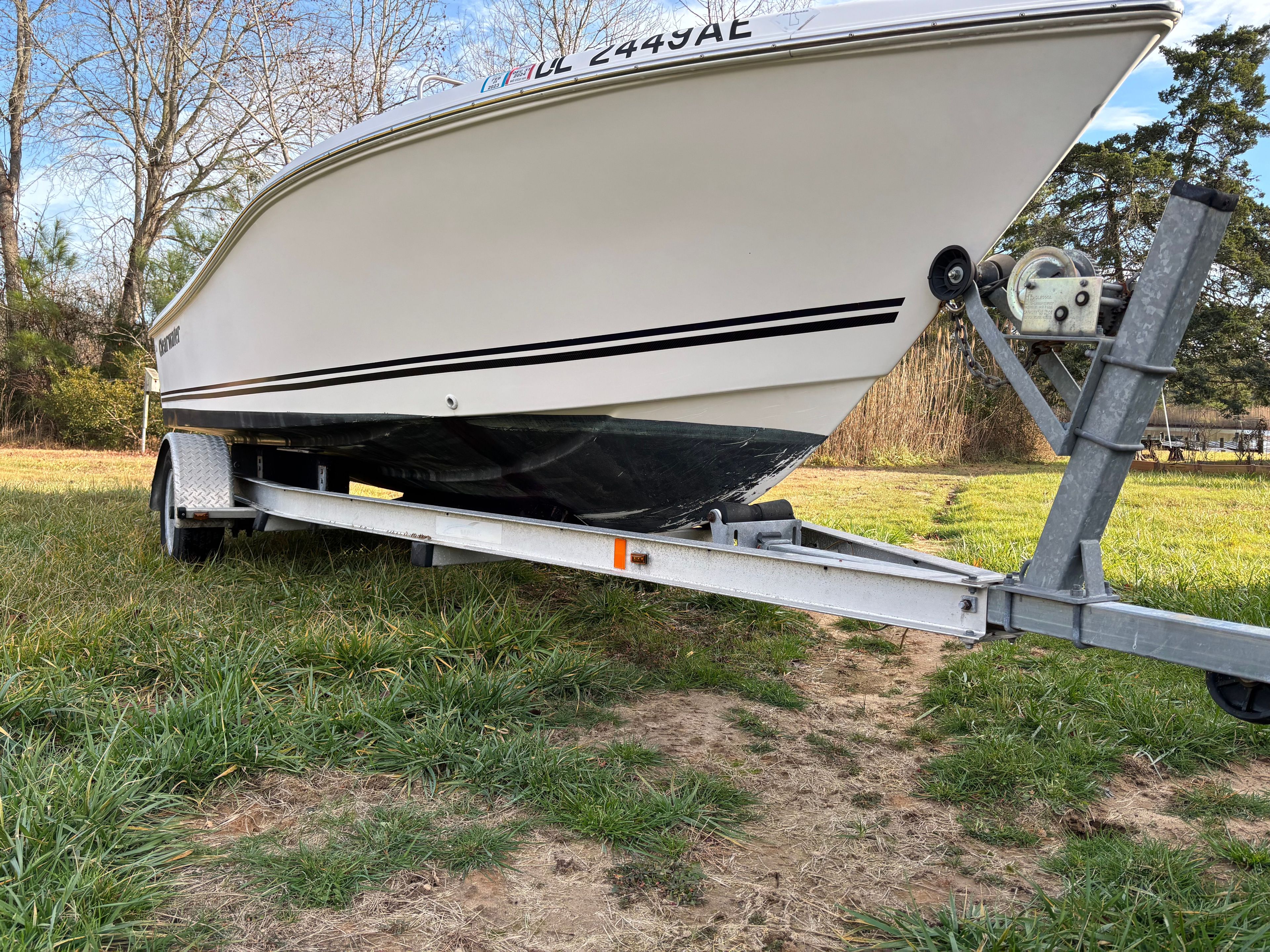 A white boat on a trailer sits on grass, surrounded by trees.