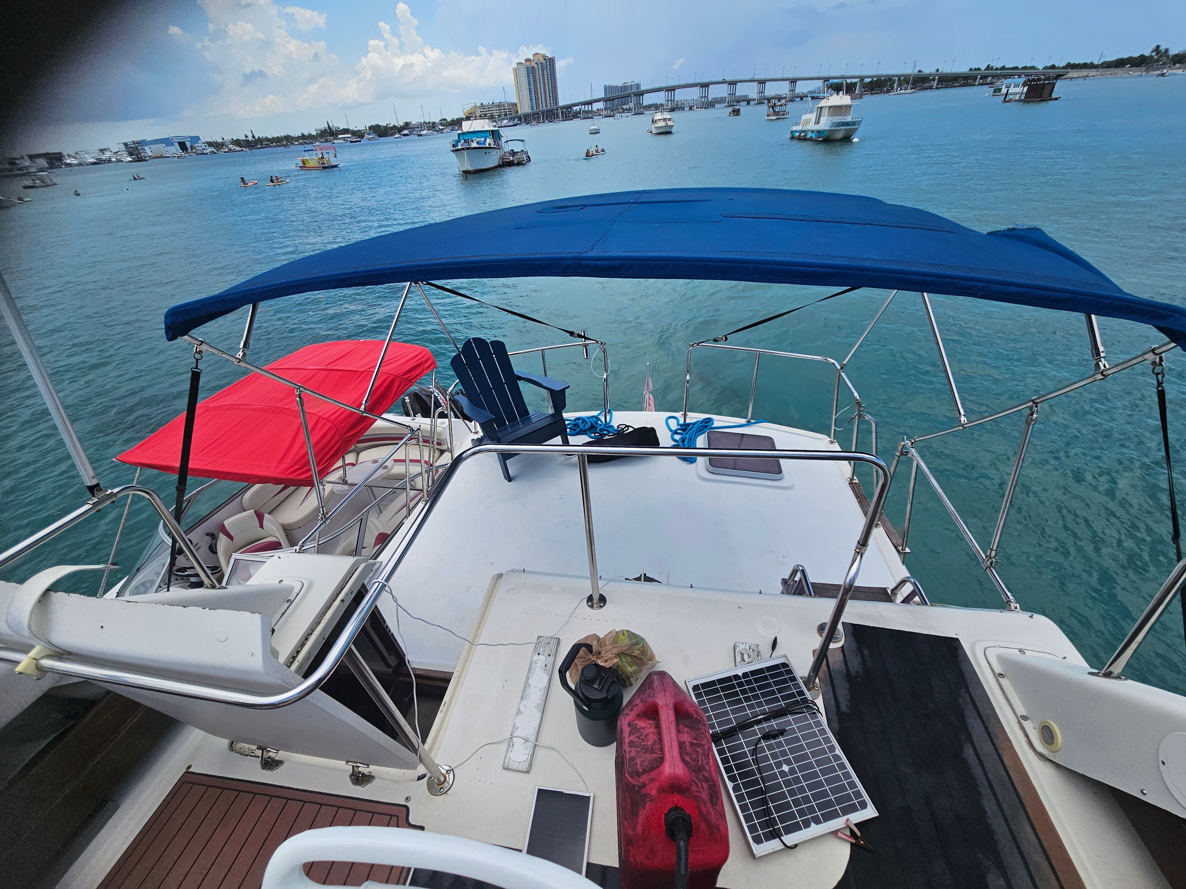 A boat with a blue sunshade on calm water, surrounded by other boats and a bridge in the background.