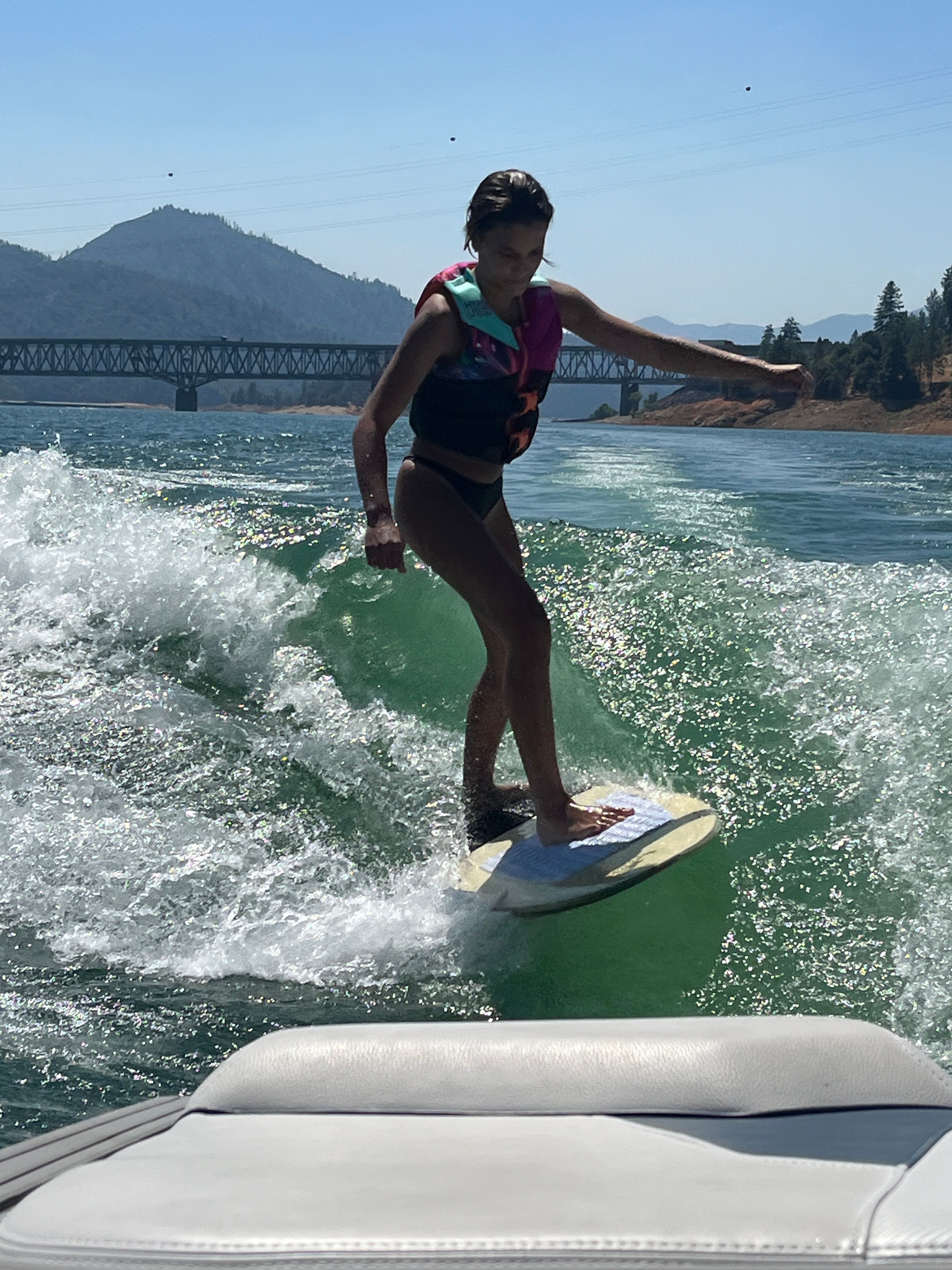 Girl surfing behind a boat on a lake with a bridge in the background.