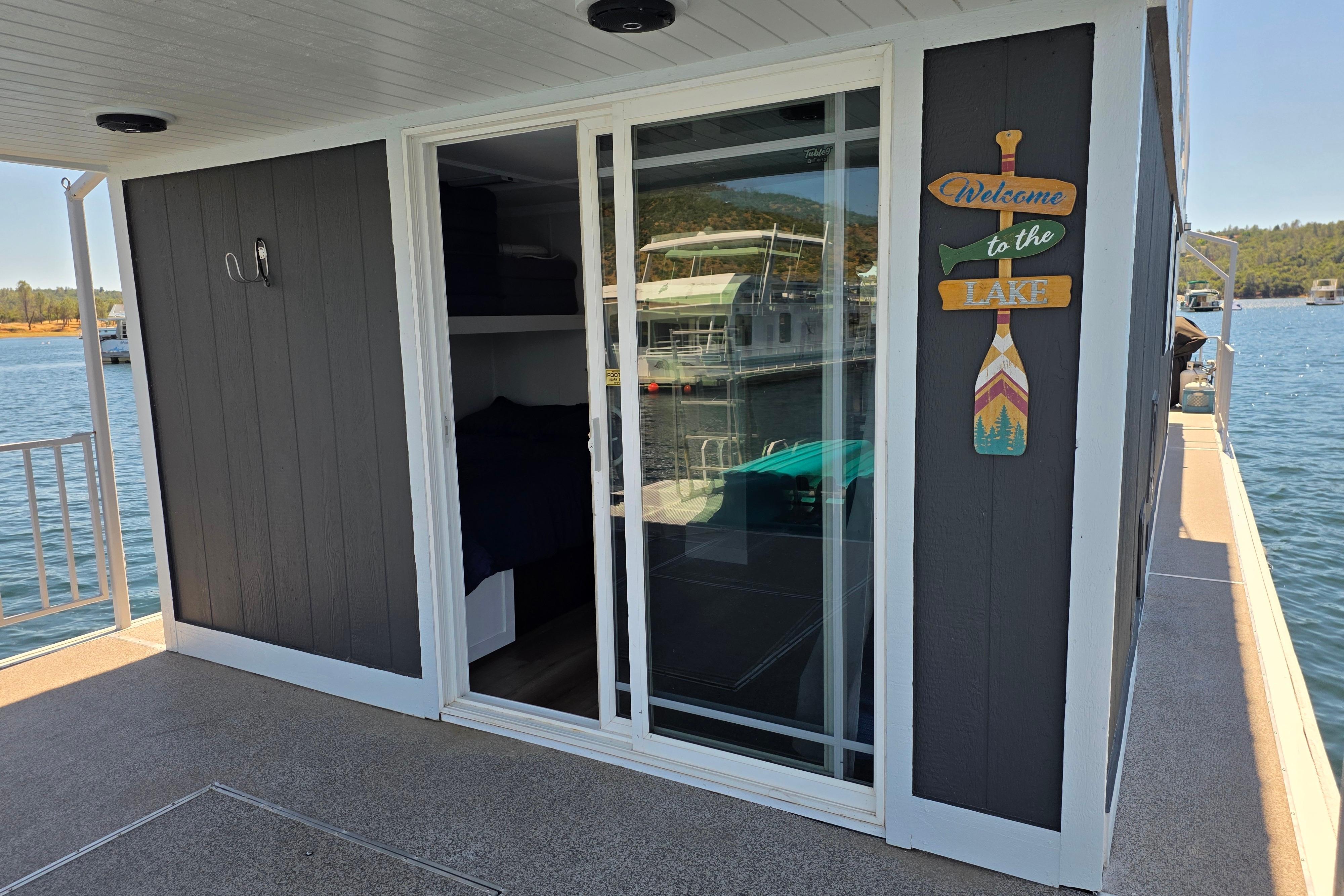 Houseboat with sliding glass door, "Welcome to the Lake" sign, docked on a sunny day.
