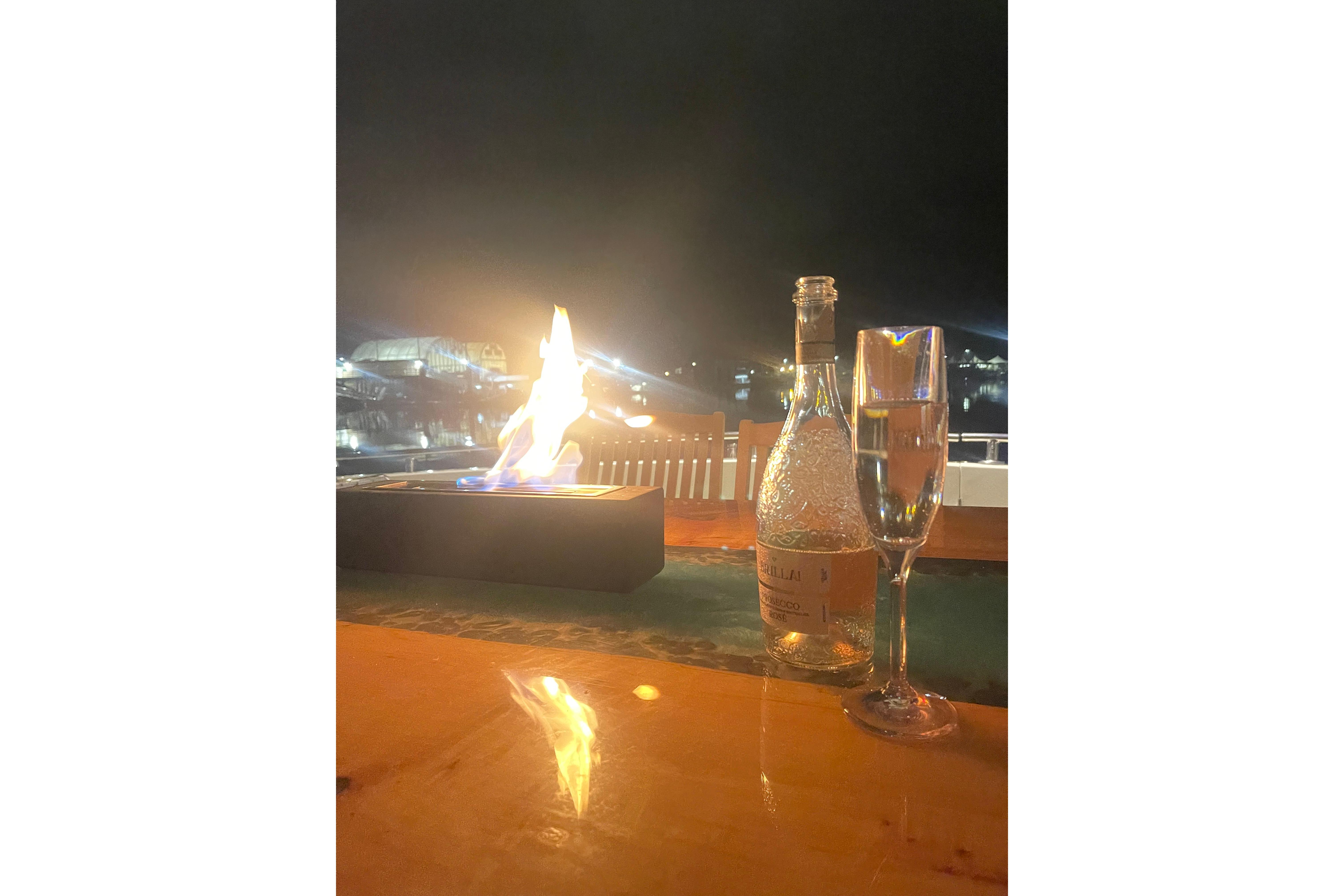 Outdoor night scene with a bottle, glass, and fire pit on a table.