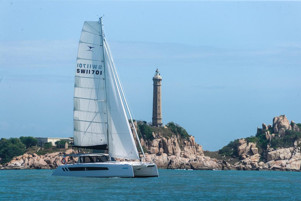 2025 Seawind 1170 catamaran sailing near rocky coastline with lighthouse in background.