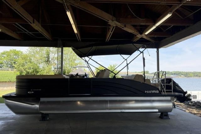 2025 Harris Sunliner 230 pontoon boat under a covered dock, near a scenic lake.