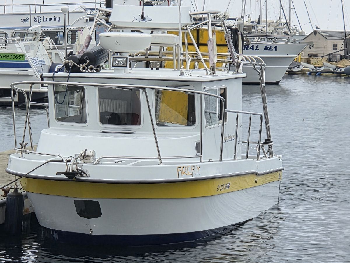 A white boat with a yellow stripe is docked in the harbor. It features a long cabin.