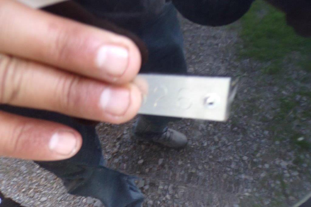 Hand holding a metal tag with engraved numbers, outdoors on gravel path.