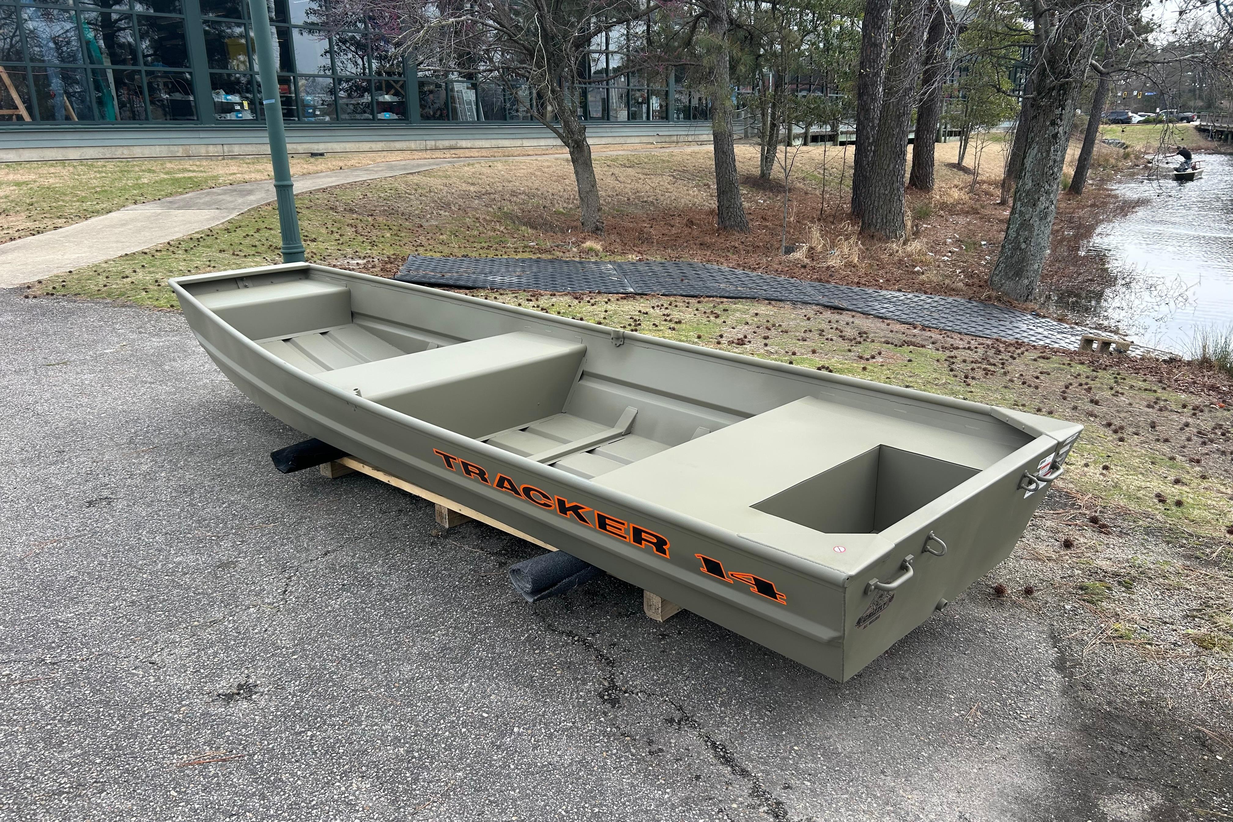 2026 Tracker Grizzly 14 Jon boat on pavement near a wooded lakeside area.