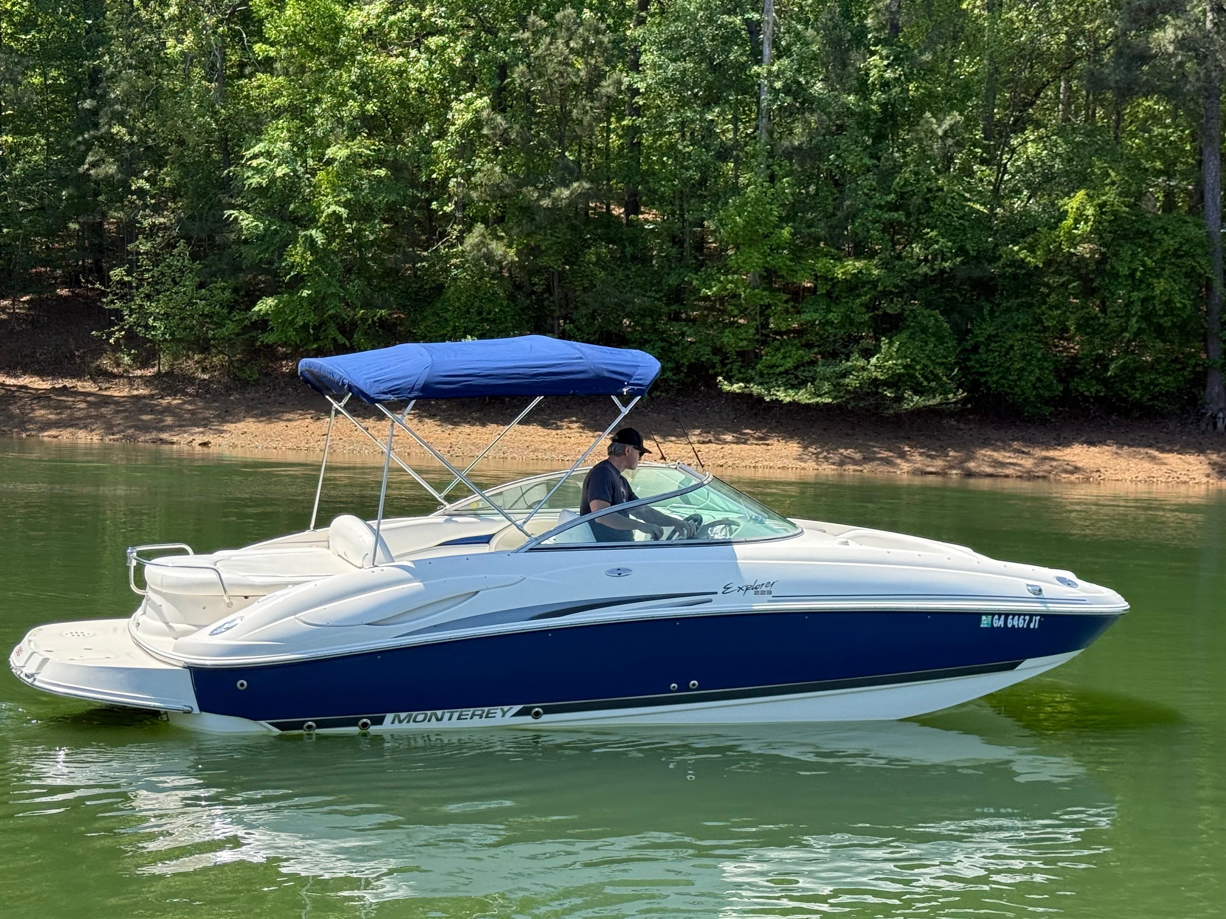 A white and blue 2005 Monterey 223 Explorer Sport boat with a sunshade on a calm lake.