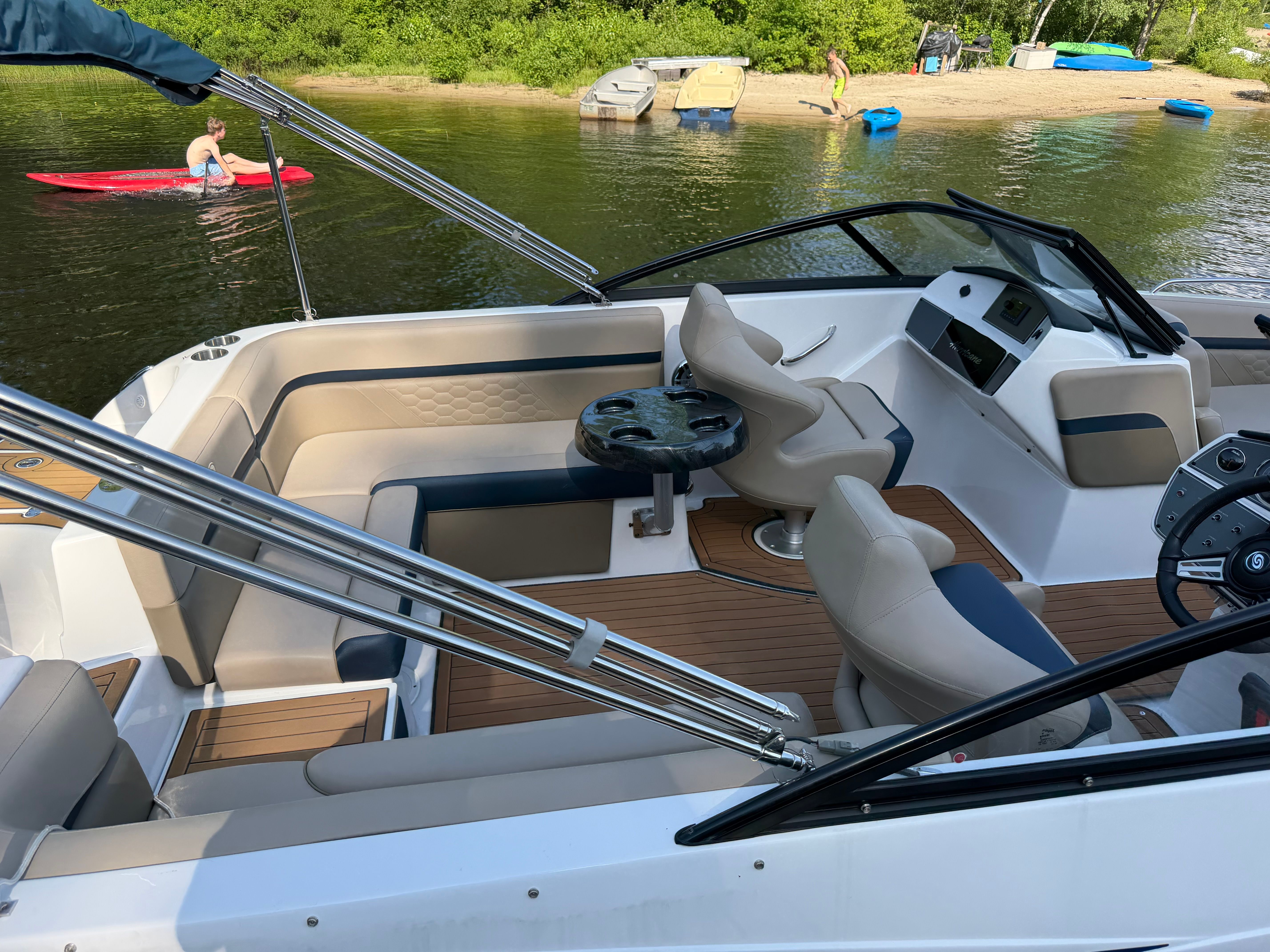 The image shows a 2022 Hurricane SunDeck 217 OB boat. It features a spacious seating area with beige and blue upholstery. A beach with a kayaker is visible in the background.