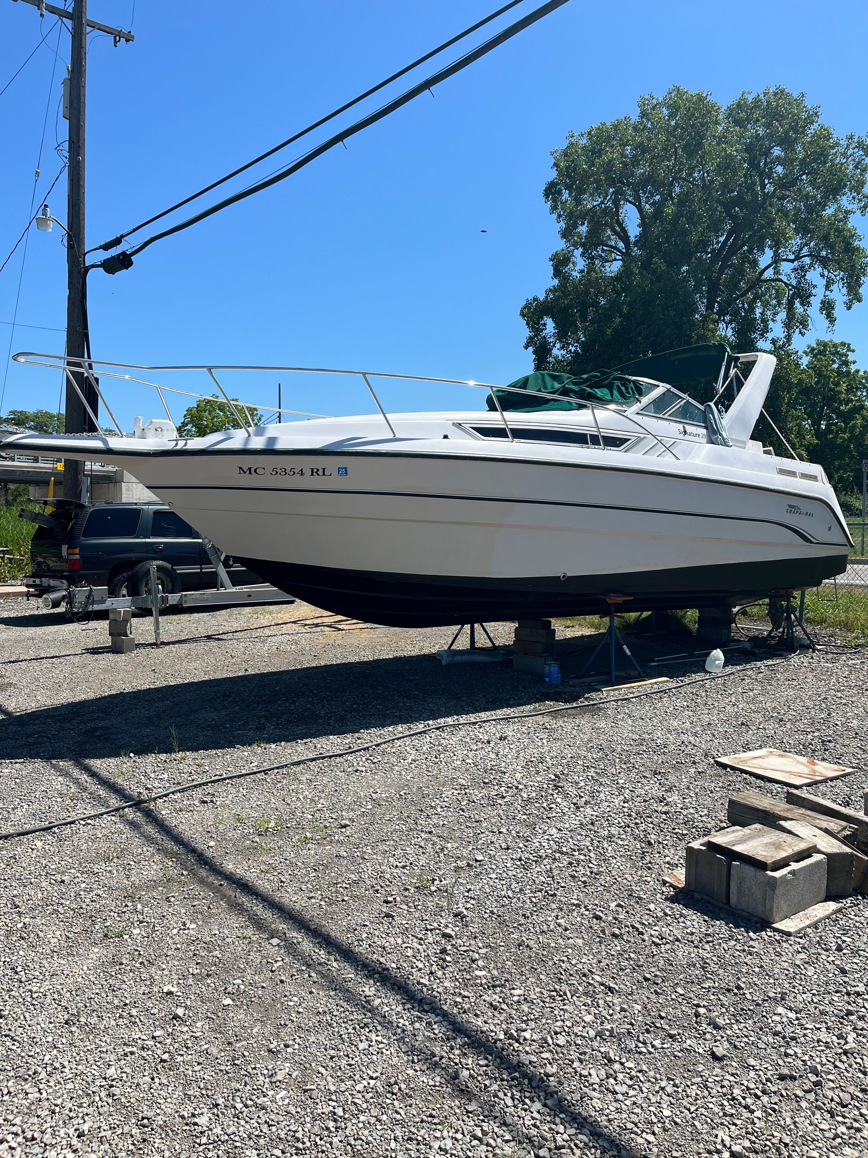 A white boat is on a gravel lot, supported by stands, with trees in the background.