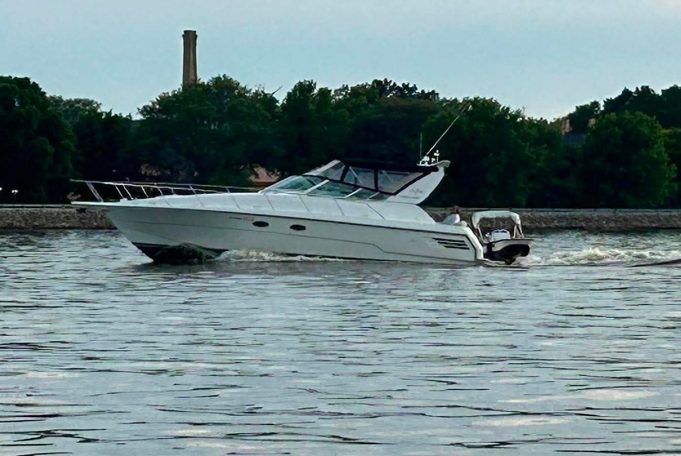 A white motorboat cruises on calm water, surrounded by trees and a chimney in the background.