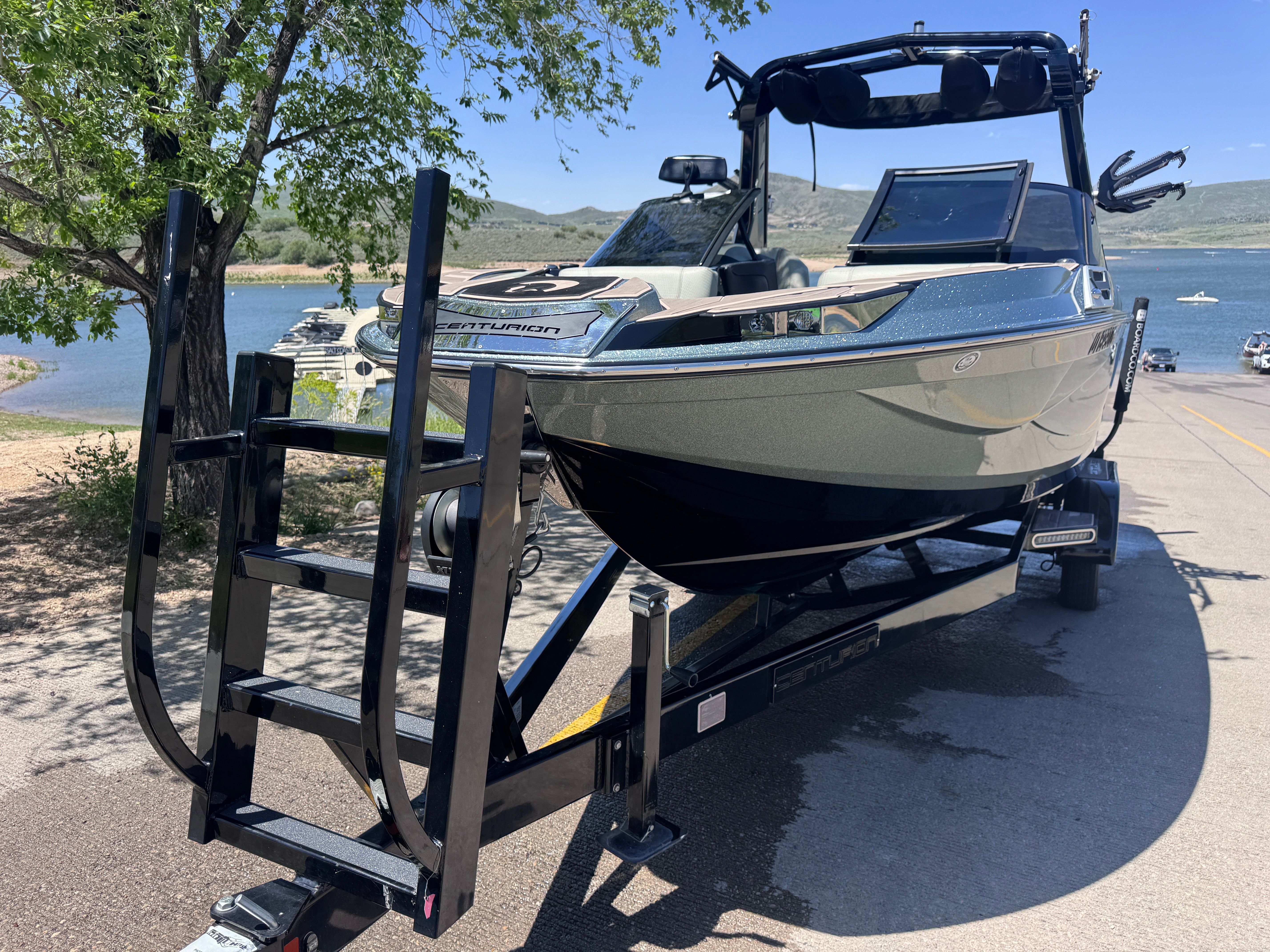 A 2022 Centurion Ri230 boat on a trailer, near a lake with hills in the background.