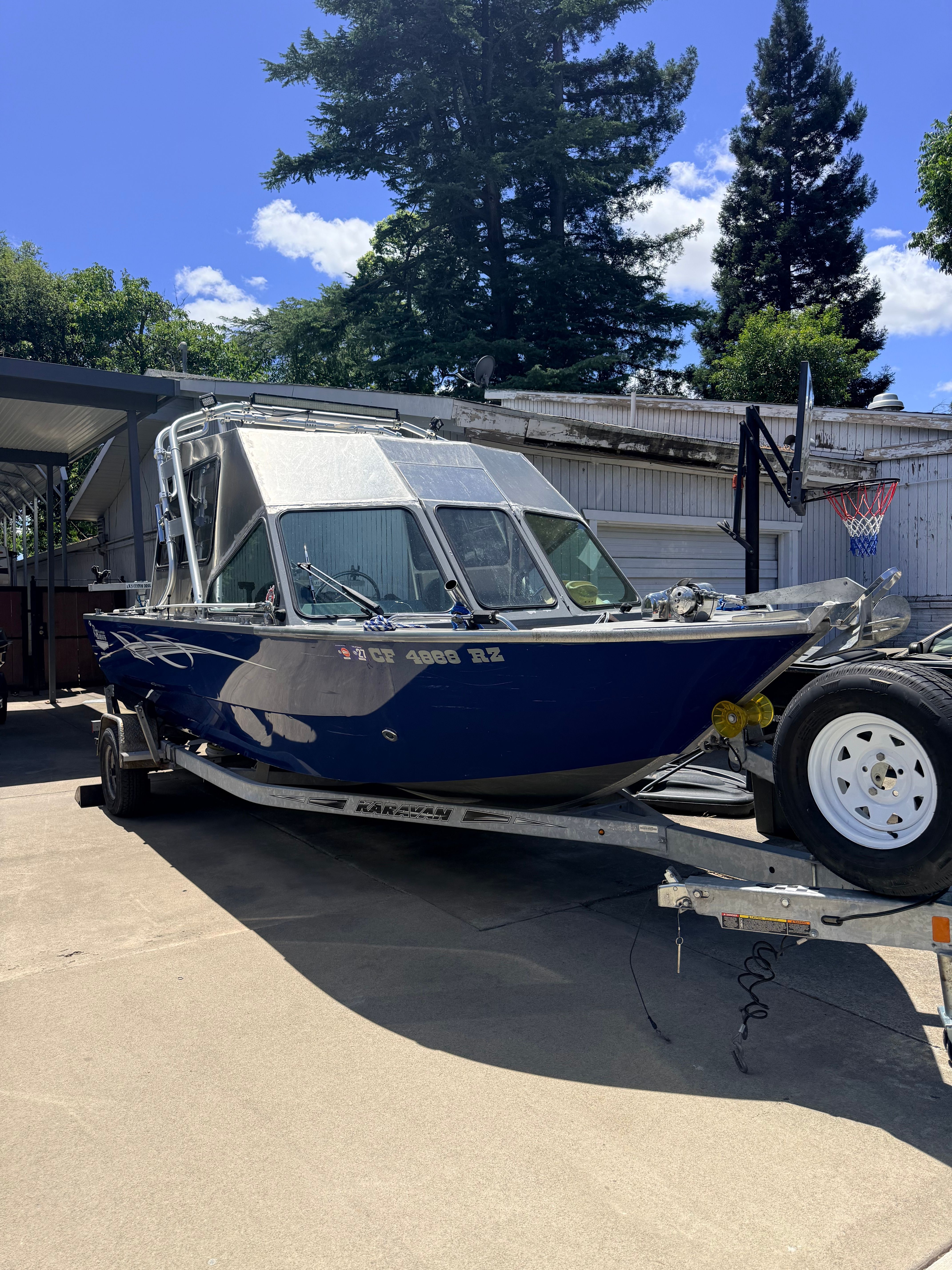 A blue River Hawk Coastal Series 20’ boat on a trailer, surrounded by trees.