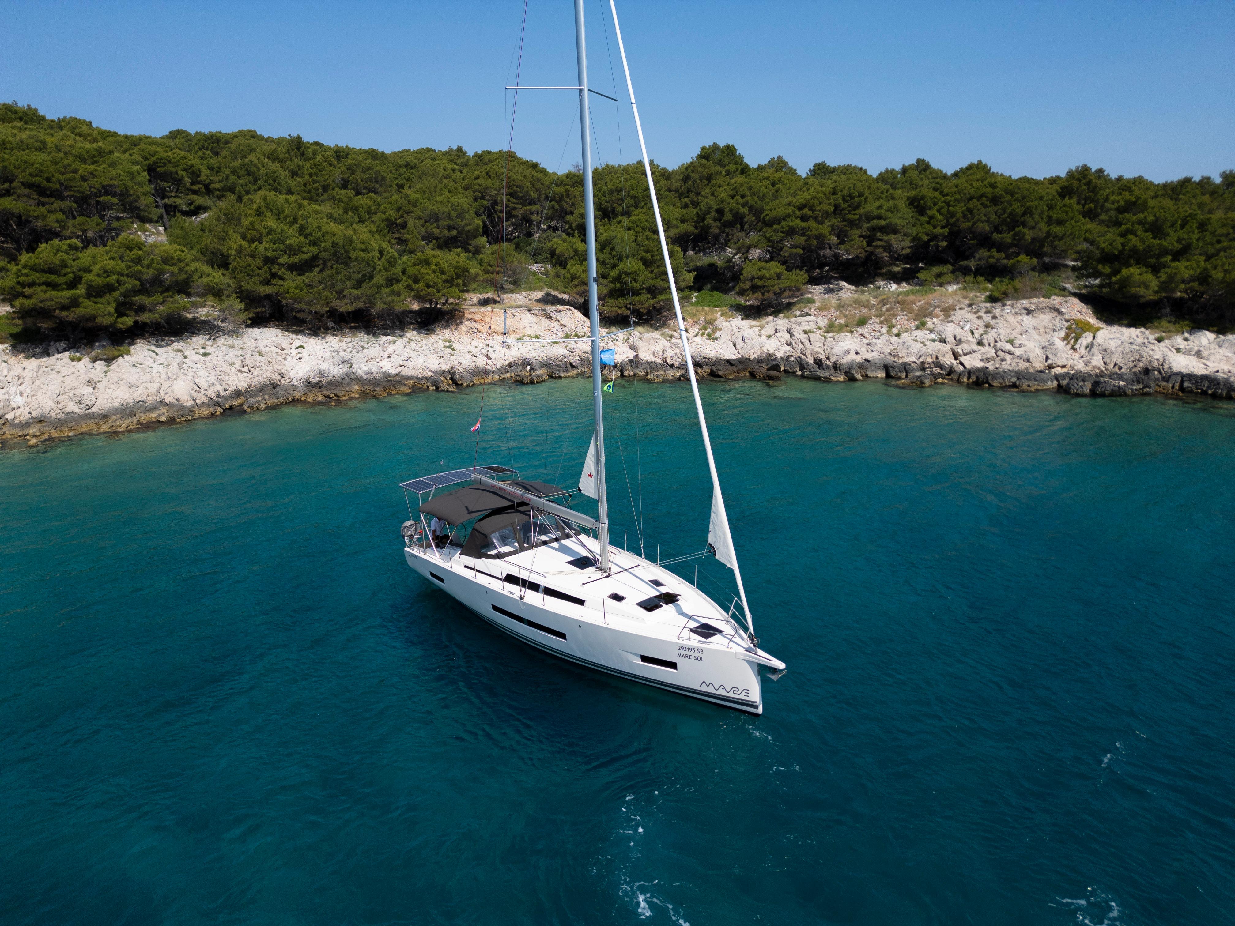Sailing yacht Hanse 410, 2025 model, anchored in clear turquoise waters near a rocky shoreline.