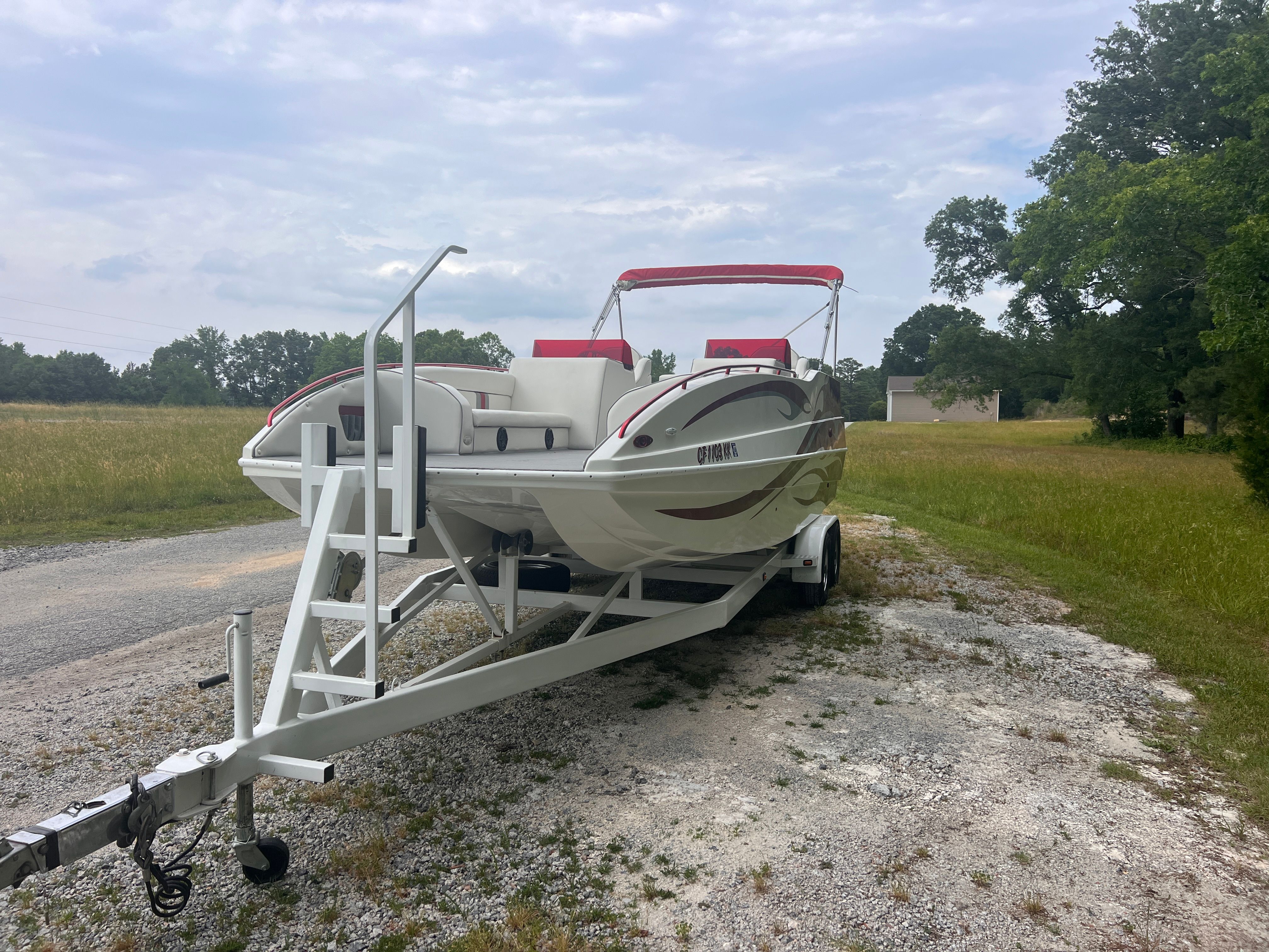 A white boat with red accents sits on a trailer on a gravel path beside a field.