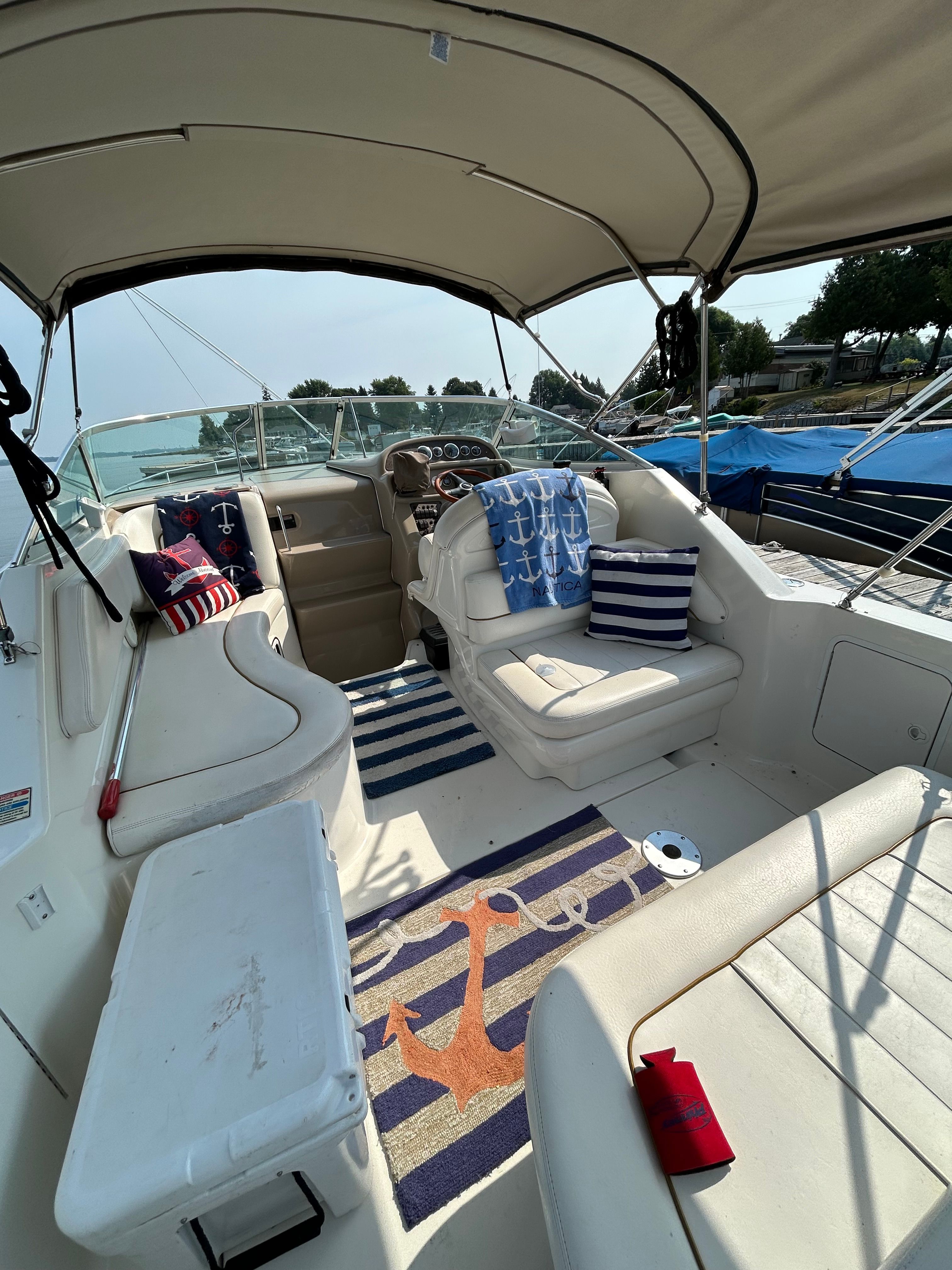 The image shows the cockpit of a 1999 Sea Ray 270 Sundancer Special Edition. There are white seats, decorative pillows with nautical themes, and a rug with an anchor design. The boat is on the water, surrounded by other boats.