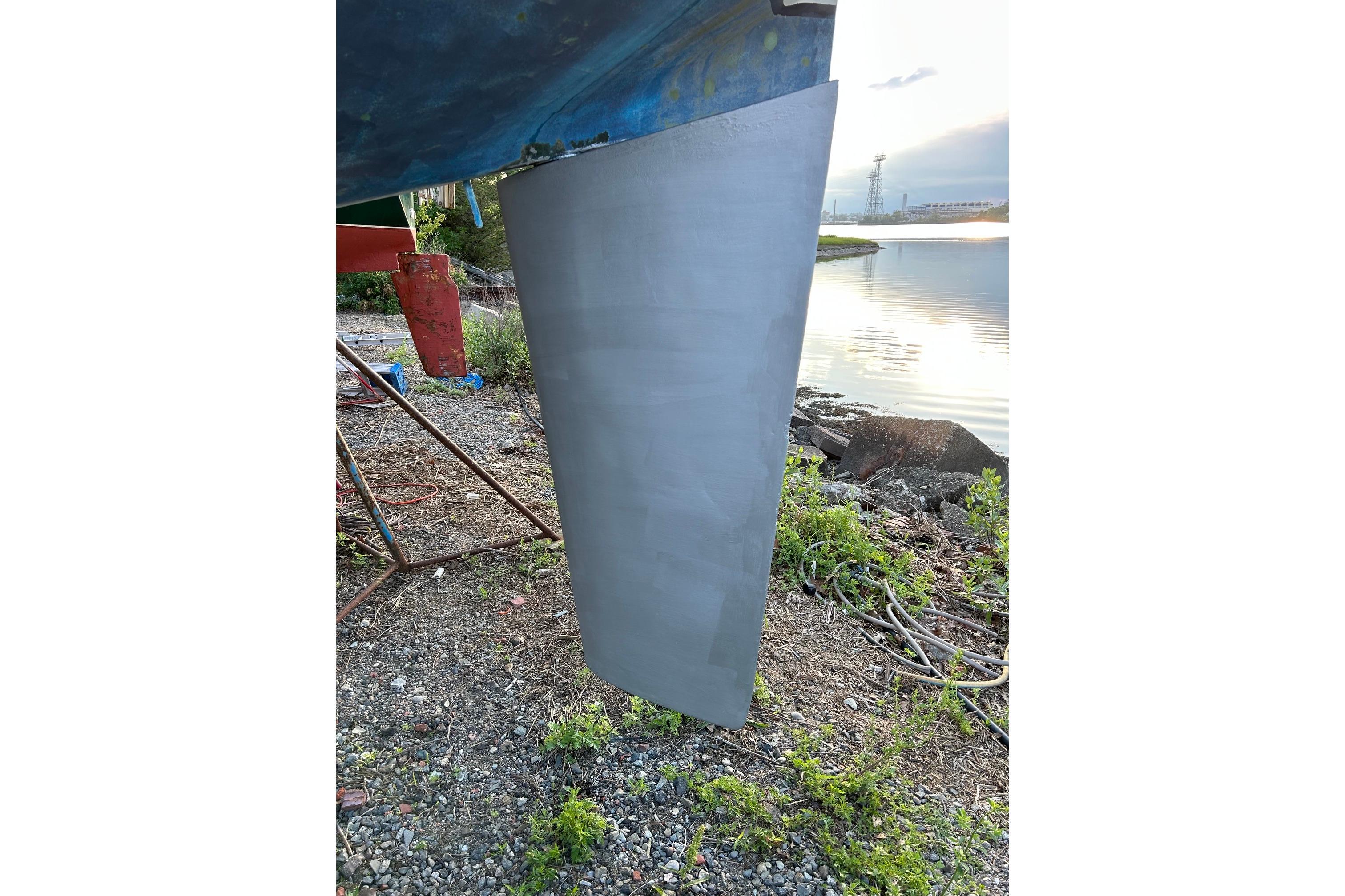 Rudder of a 1987 Ericson 38-200 sailboat on a gravel shore near water.