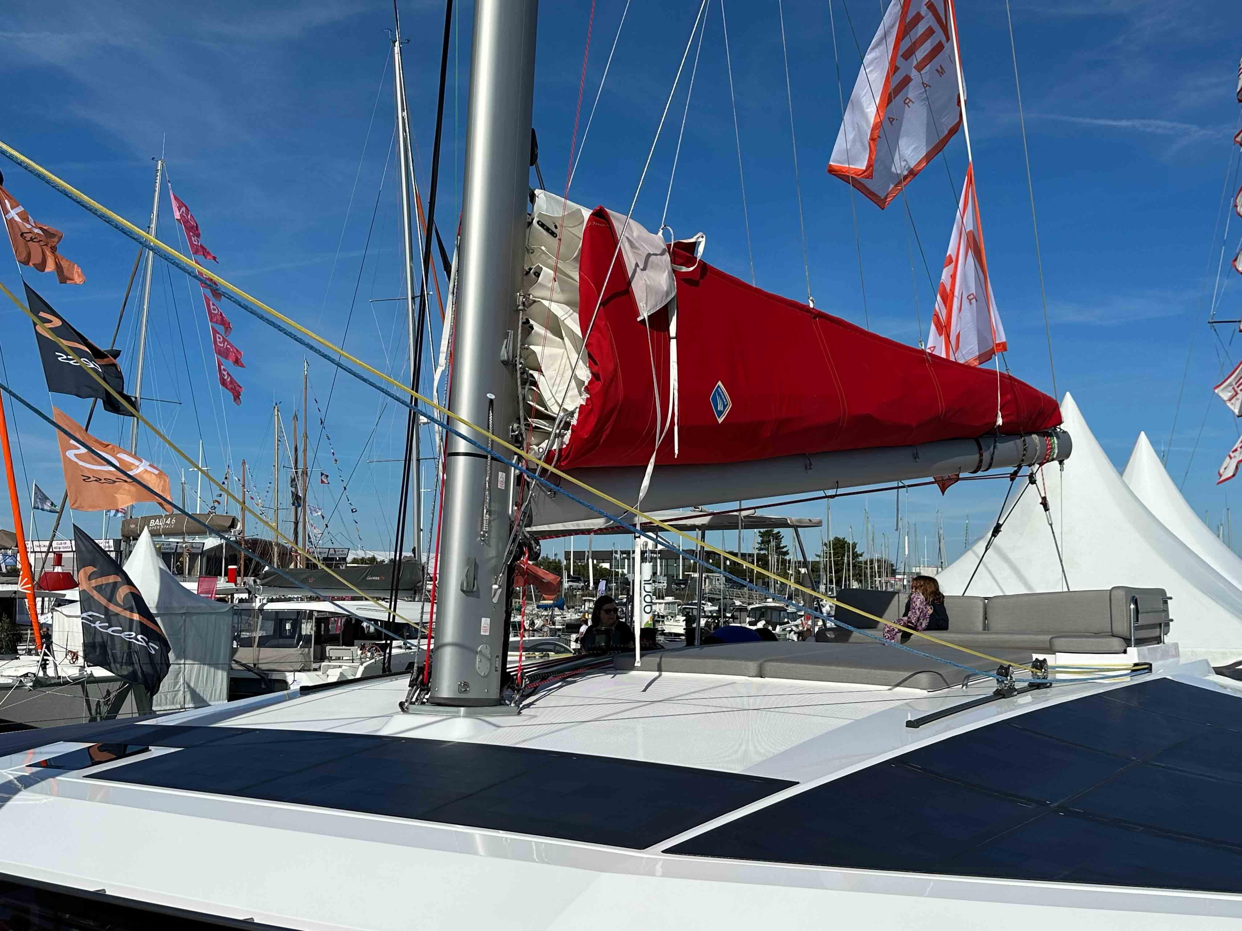 Sailing yacht NEEL 52 Hybrid 2025 with red sail at marina, surrounded by flags.