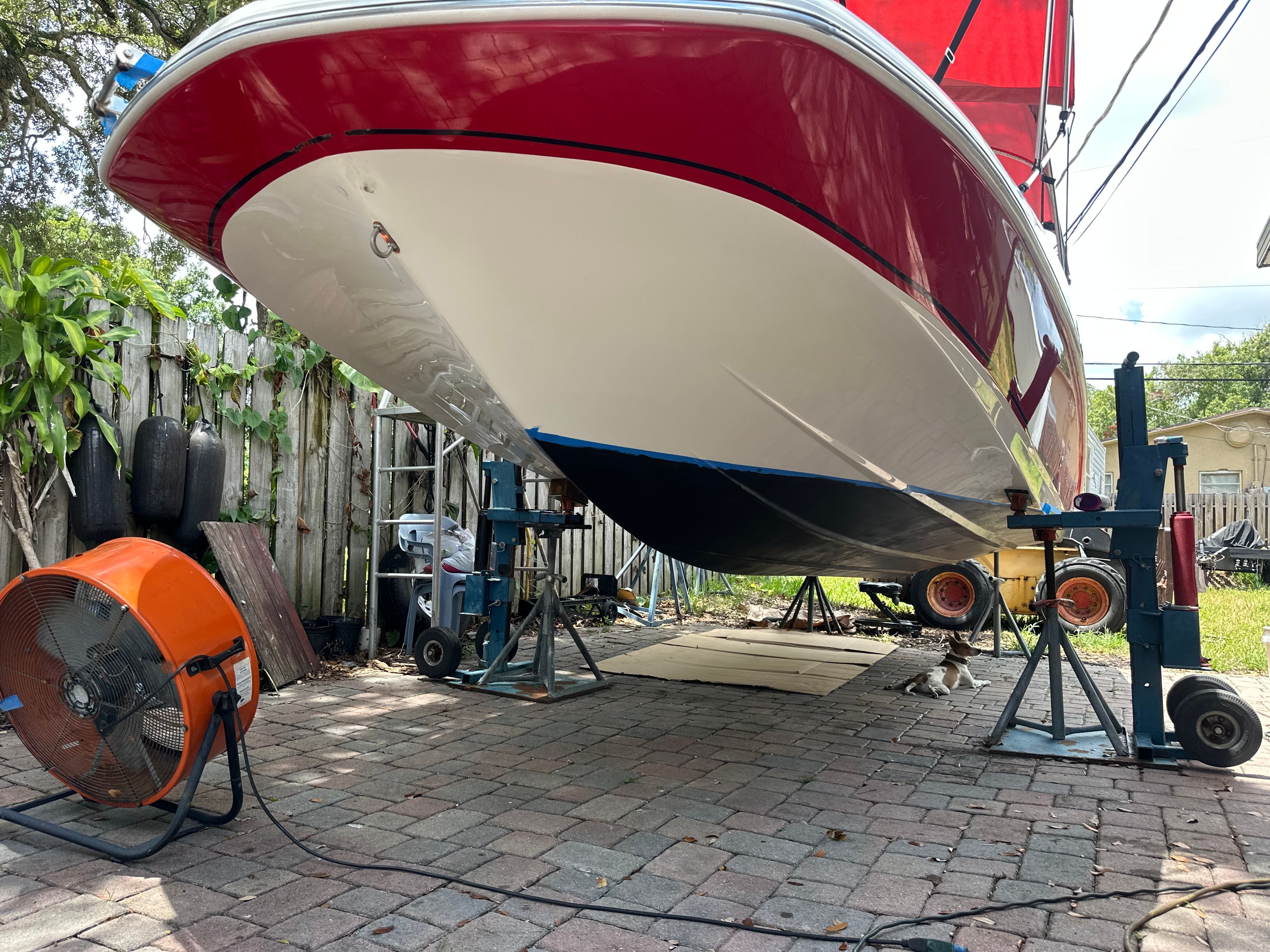 A red boat is on a paved yard, supported by jacks. A fan is nearby.