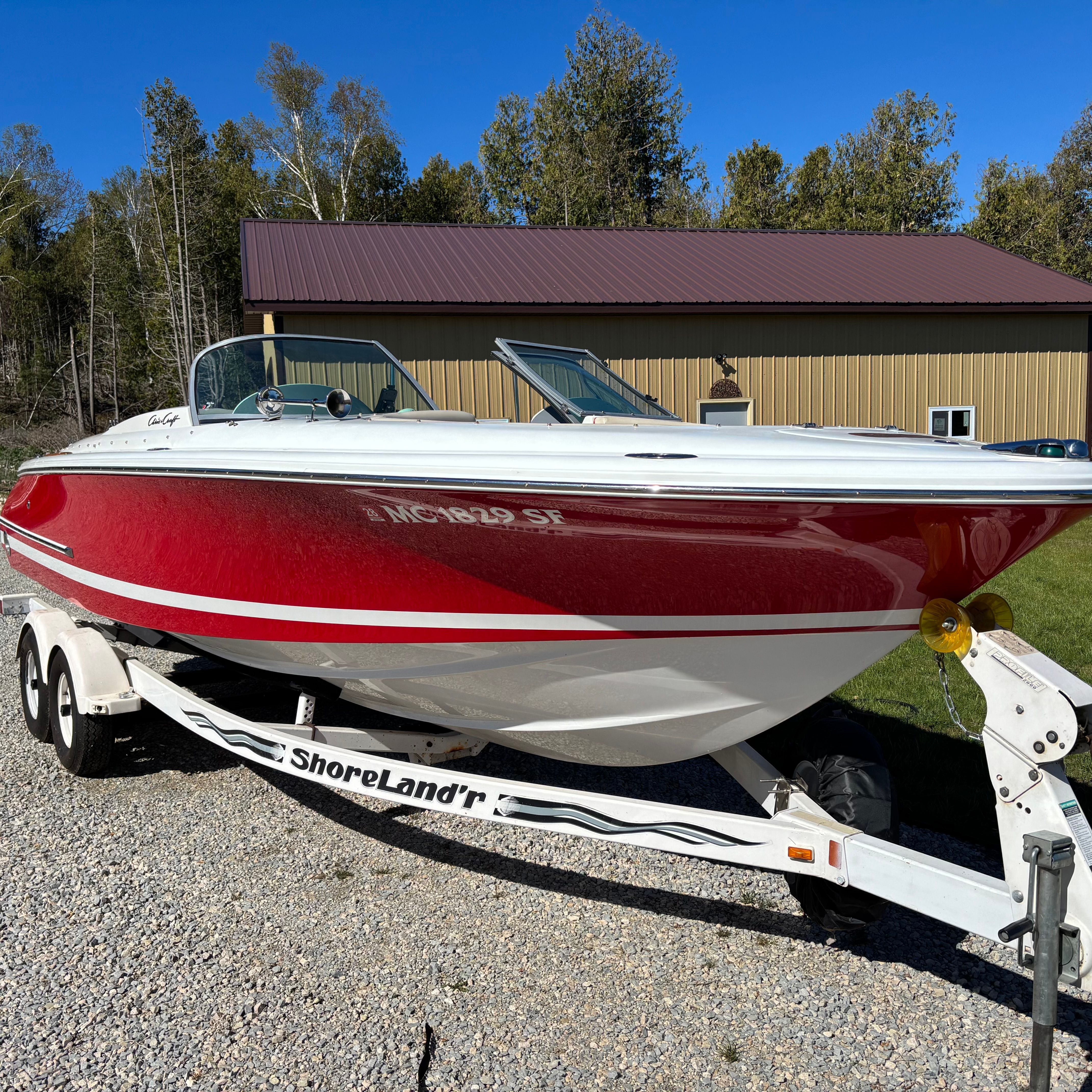 A red 2003 Chris-Craft Launch 22 boat on a trailer, surrounded by trees and a building.