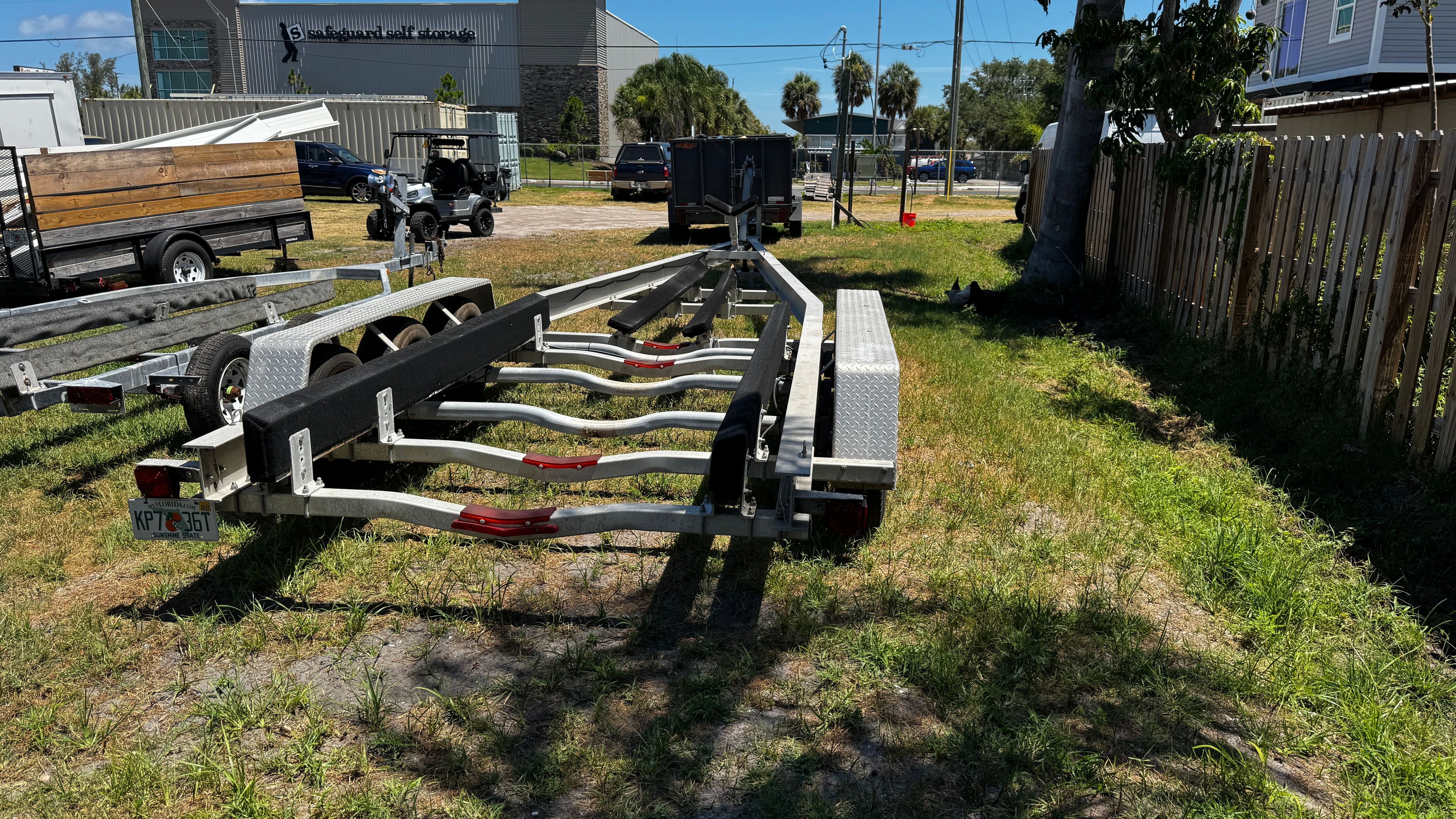 Boat trailer on grassy area next to wooden fence and trees.