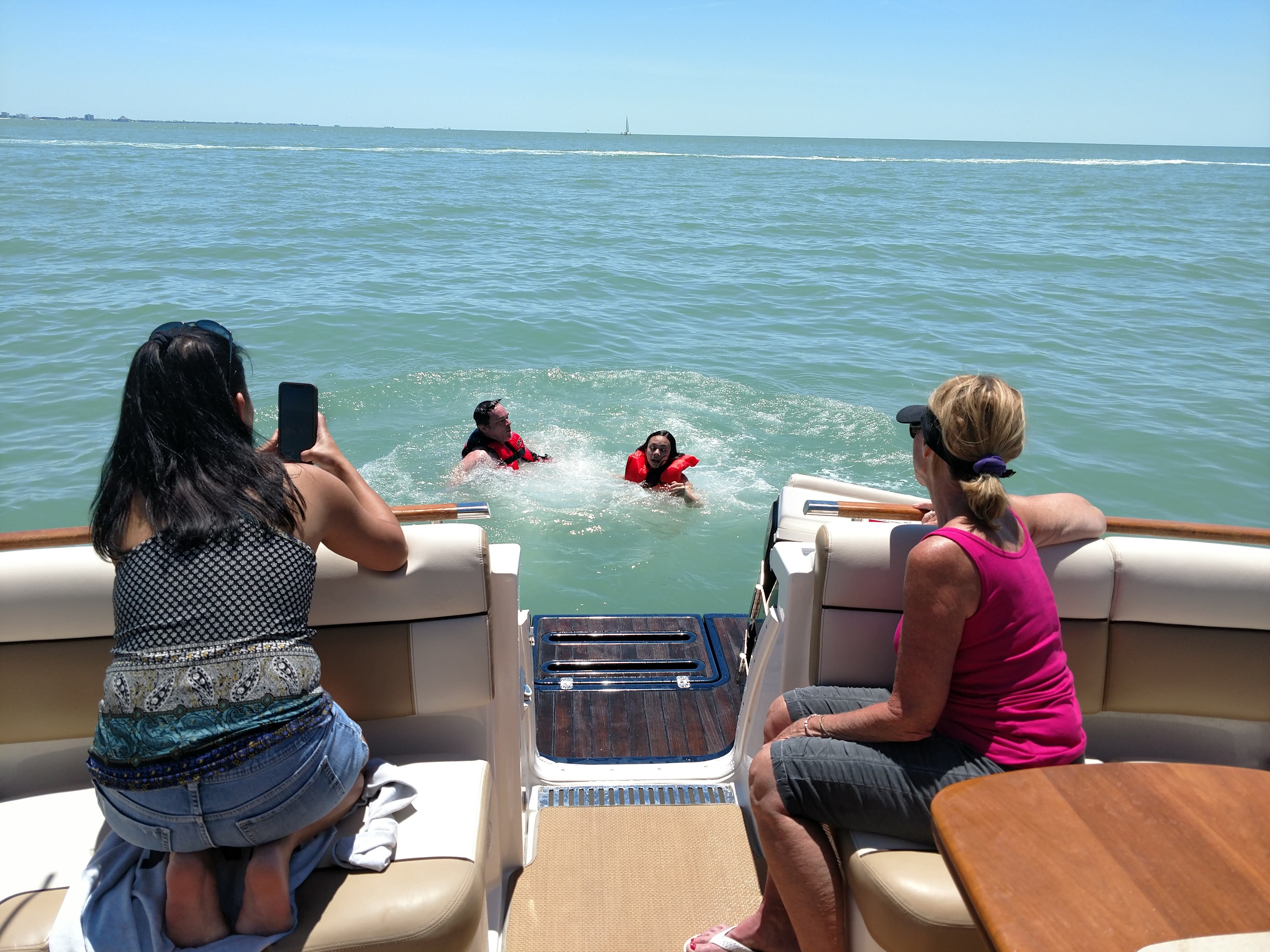 Two people swim in the water behind a yacht, while others watch from onboard.