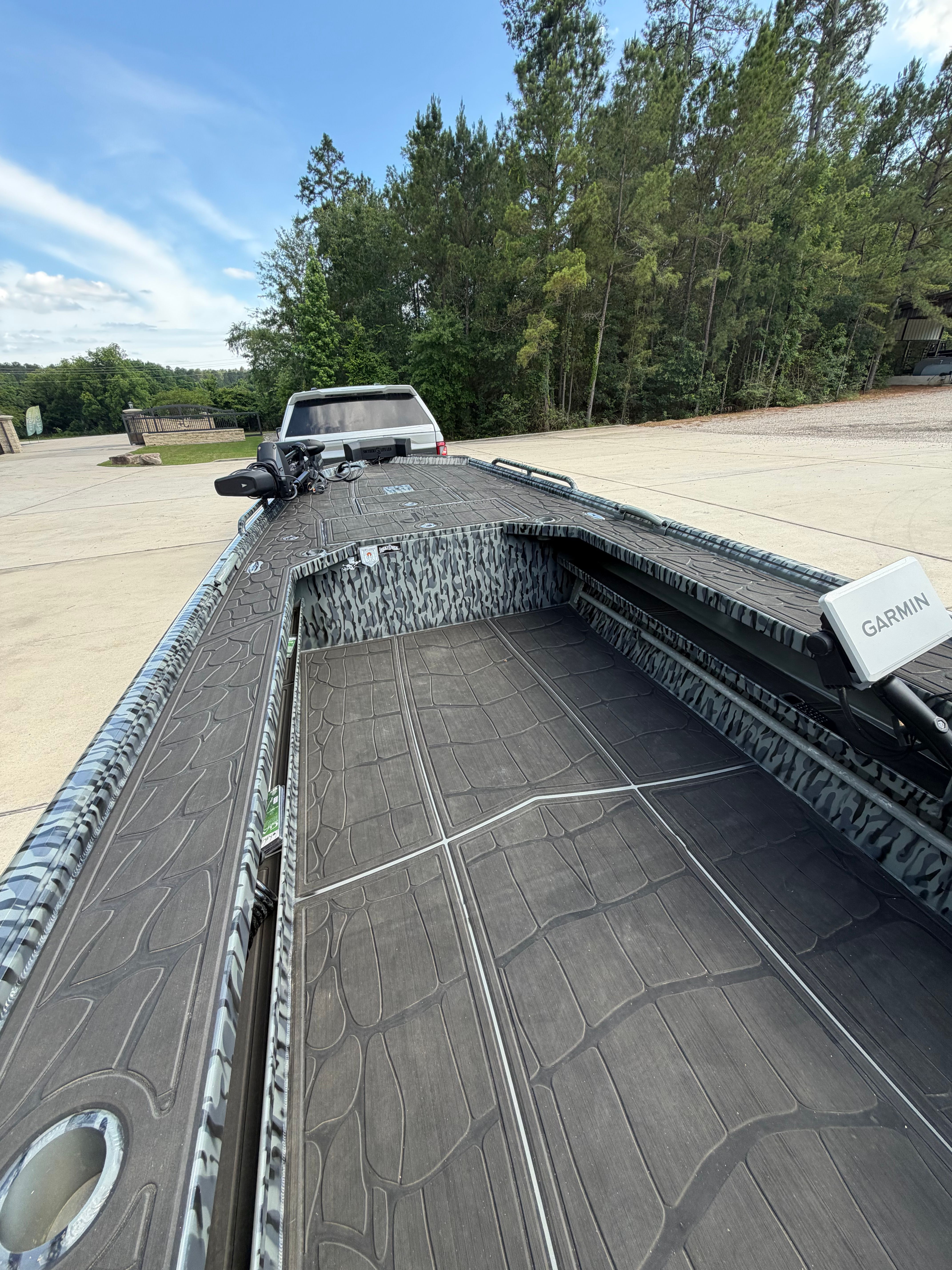A modern boat with camouflage pattern sits on a trailer in front of a forest.