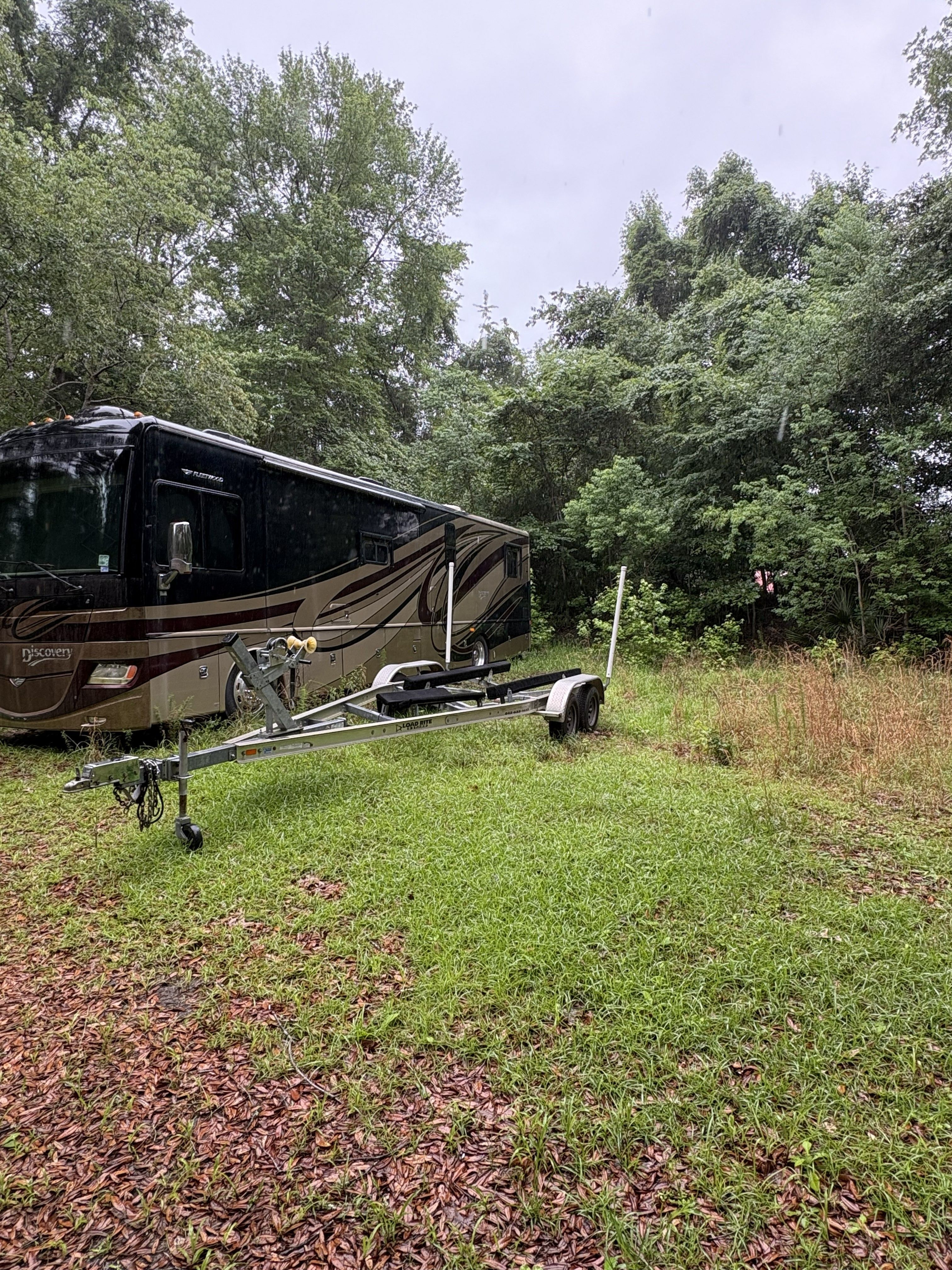 RV and boat trailer on grassy area surrounded by trees.