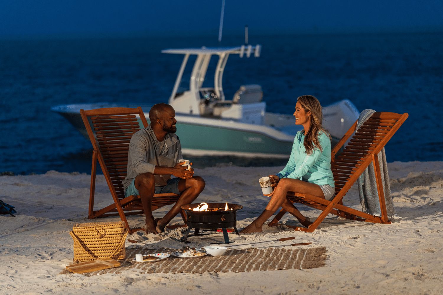 A couple sits on the beach by a campfire, with a boat in the background on the water.