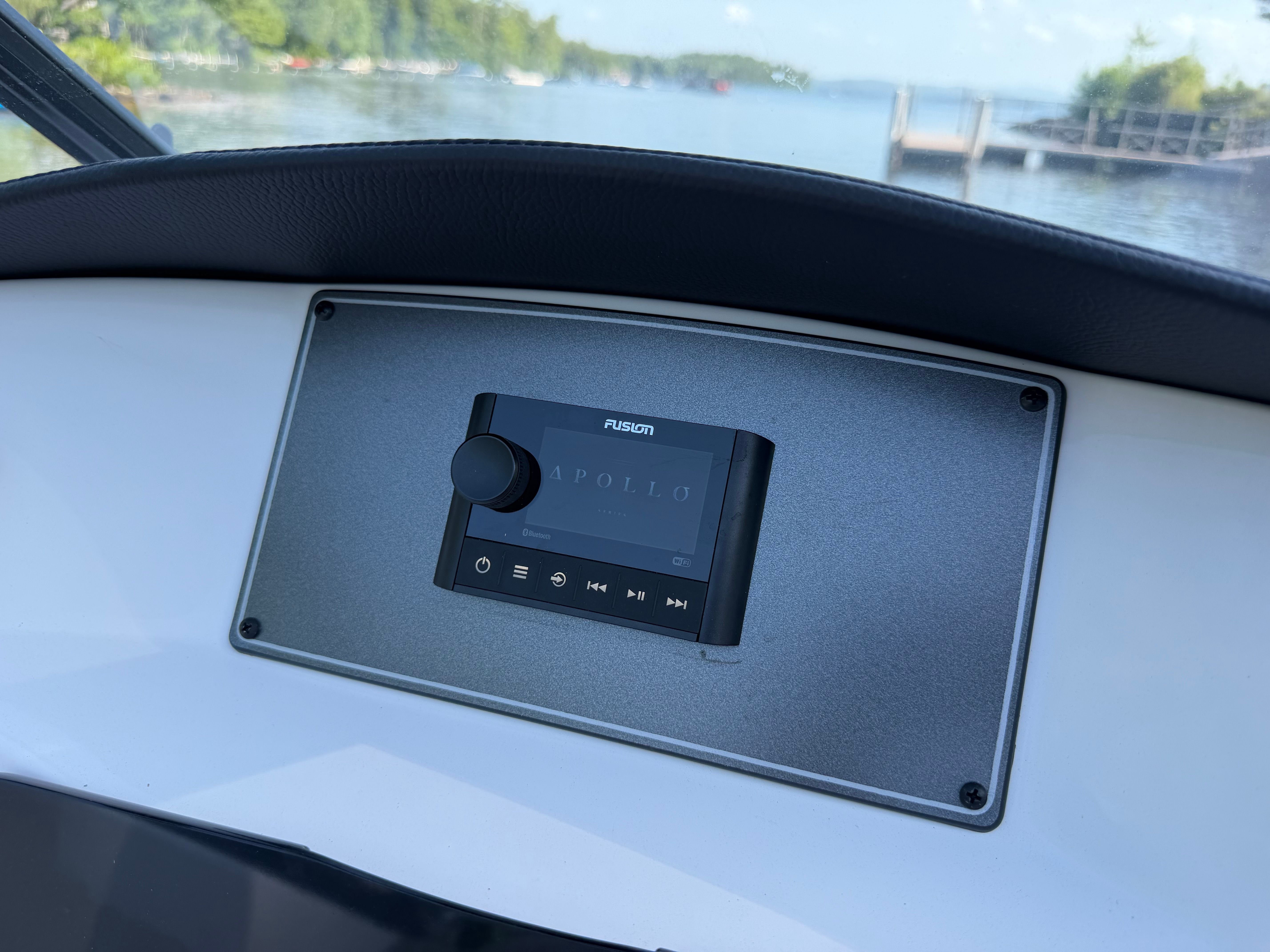 The image shows a Fusion Apollo audio system on a boat panel overlooking a lake.