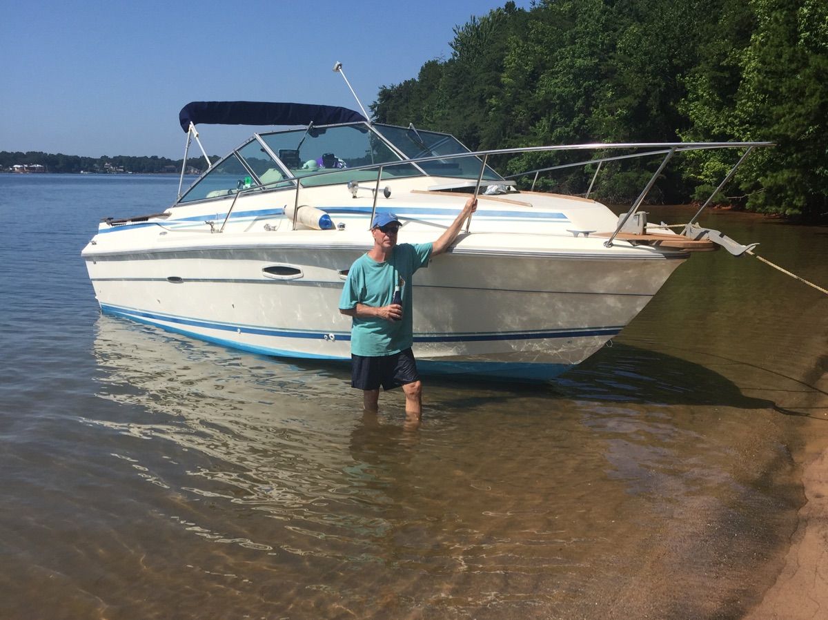 A man stands beside a Sea Ray Amberjack boat in shallow water near the shore.