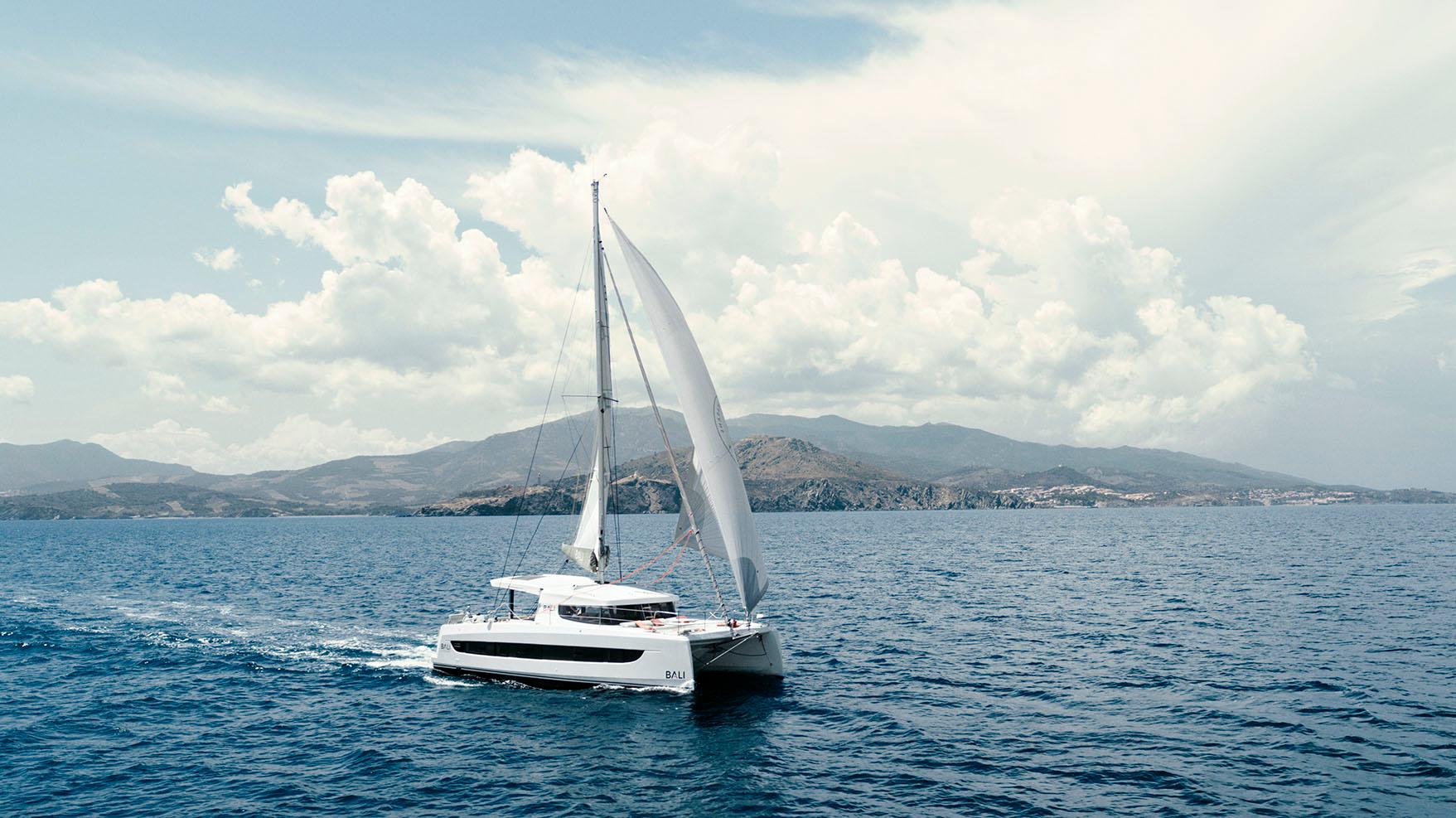 Sailing catamaran Bali CATSMART 2026 on open sea with scenic mountain backdrop.