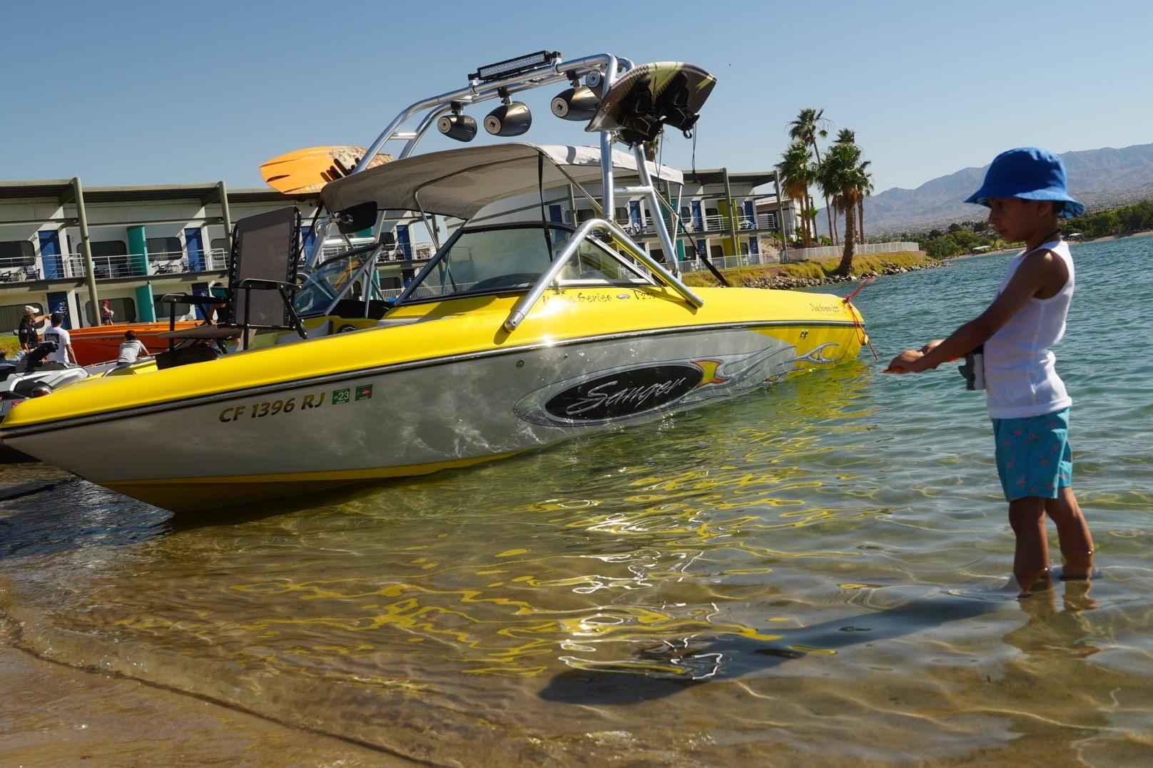 Child playing near a 2006 Sanger V230 boat on a sunny lakeside.
