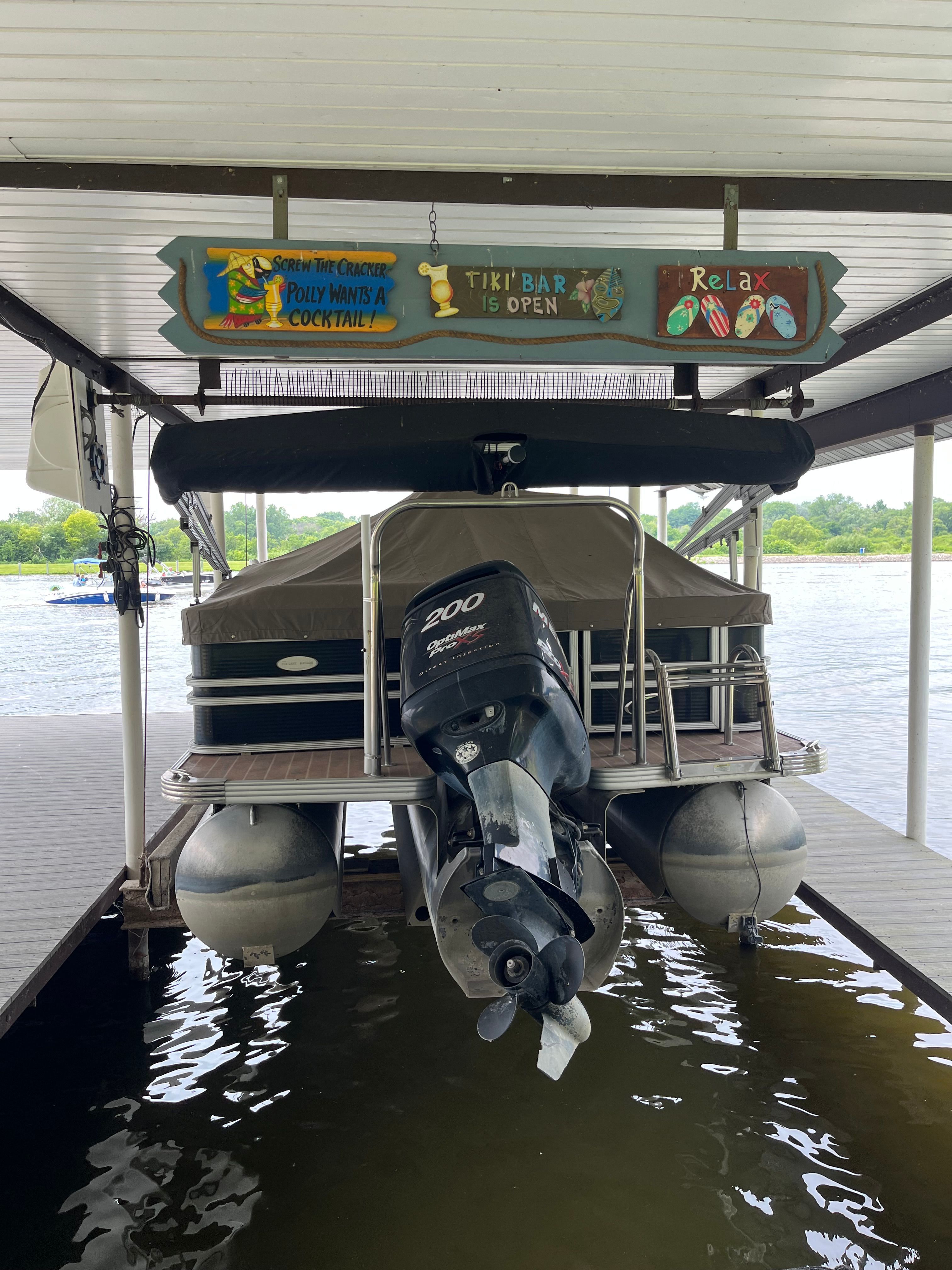 A 2012 Crest CARRIBEAN pontoon boat with an outboard motor, under a covered dock.