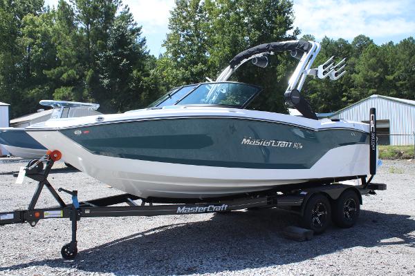 2025 MasterCraft NXT21 boat on trailer, parked outdoors, surrounded by trees and buildings.