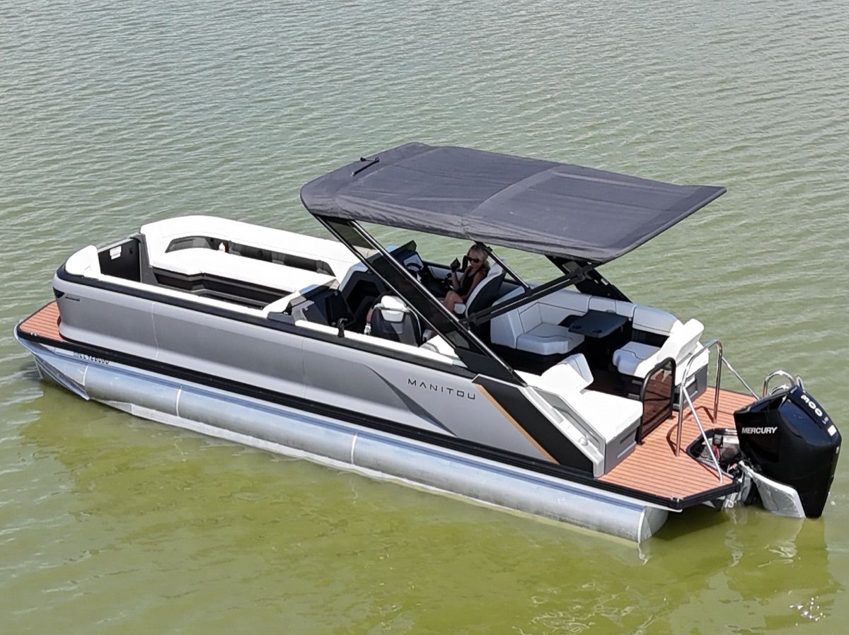 A modern pontoon boat by Manitou, Explore model 2024, with a sunshade on calm water.